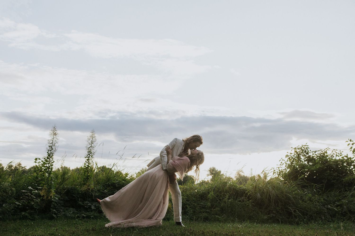A romantic couple embraces in a flowing beige dress against a cloudy sky and green natural landscape at dusk.