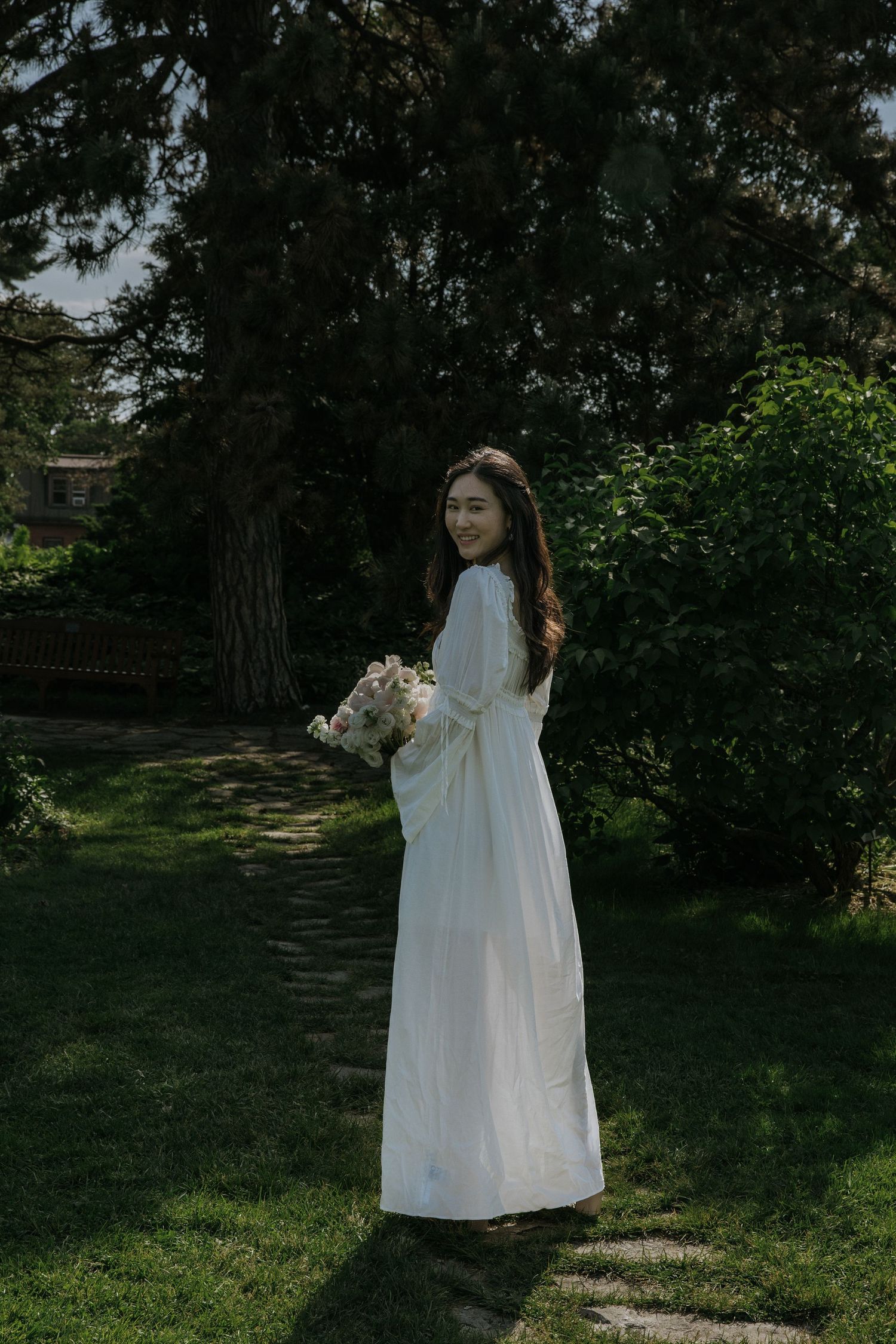A figure in a flowing white dress walks through a sunlit garden surrounded by lush greenery and trees.