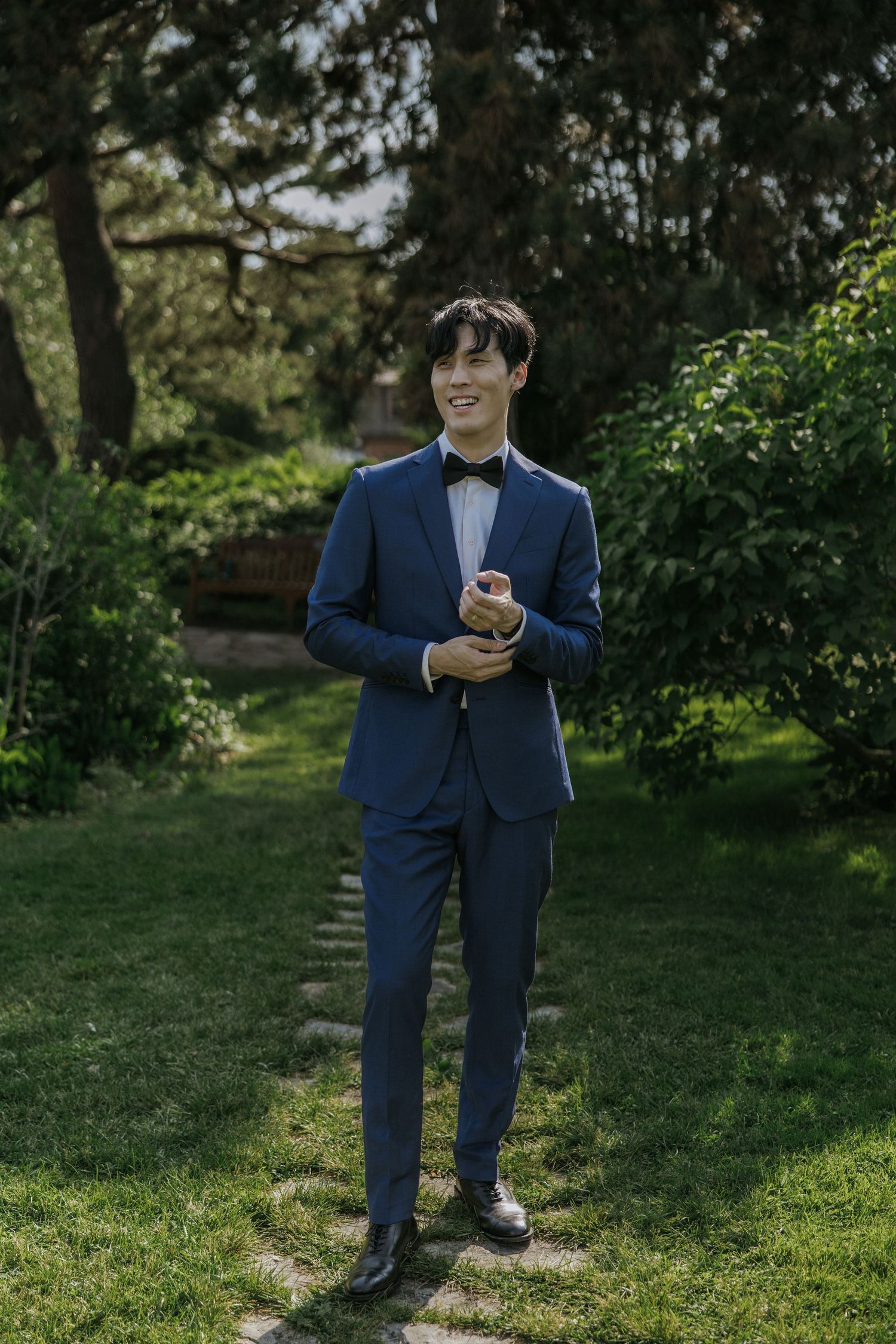 A person in a navy blue suit stands on grass in a garden setting, looking at their mobile phone.