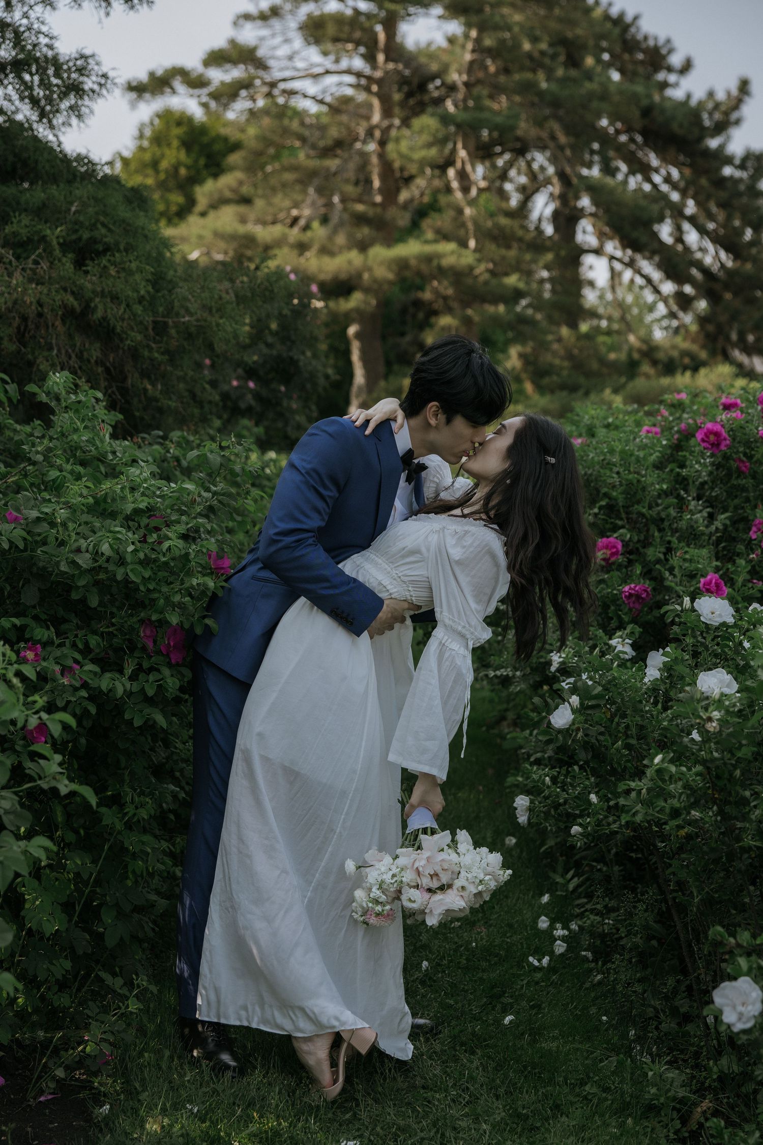 A romantic couple embraces in a lush garden setting with blooming flowers and trees in the background.