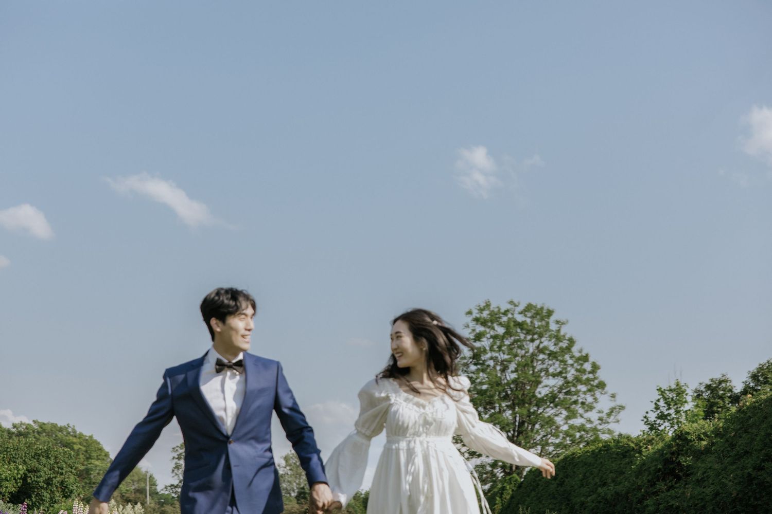 A couple in wedding attire walks hand in hand through a lush green garden setting on their special day.