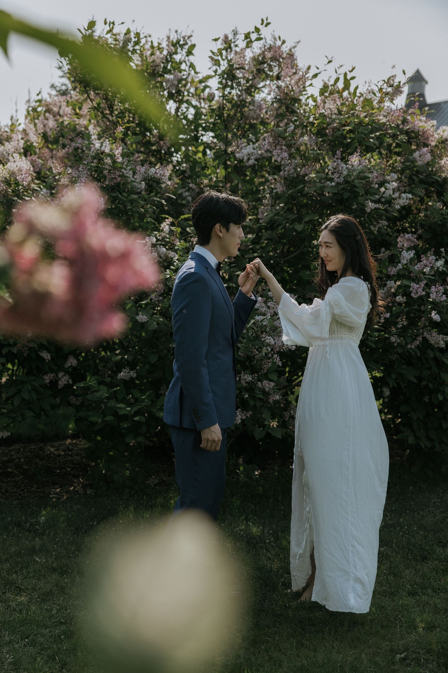 A couple in wedding attire stands together in a garden setting with blooming trees and flowers in the background.