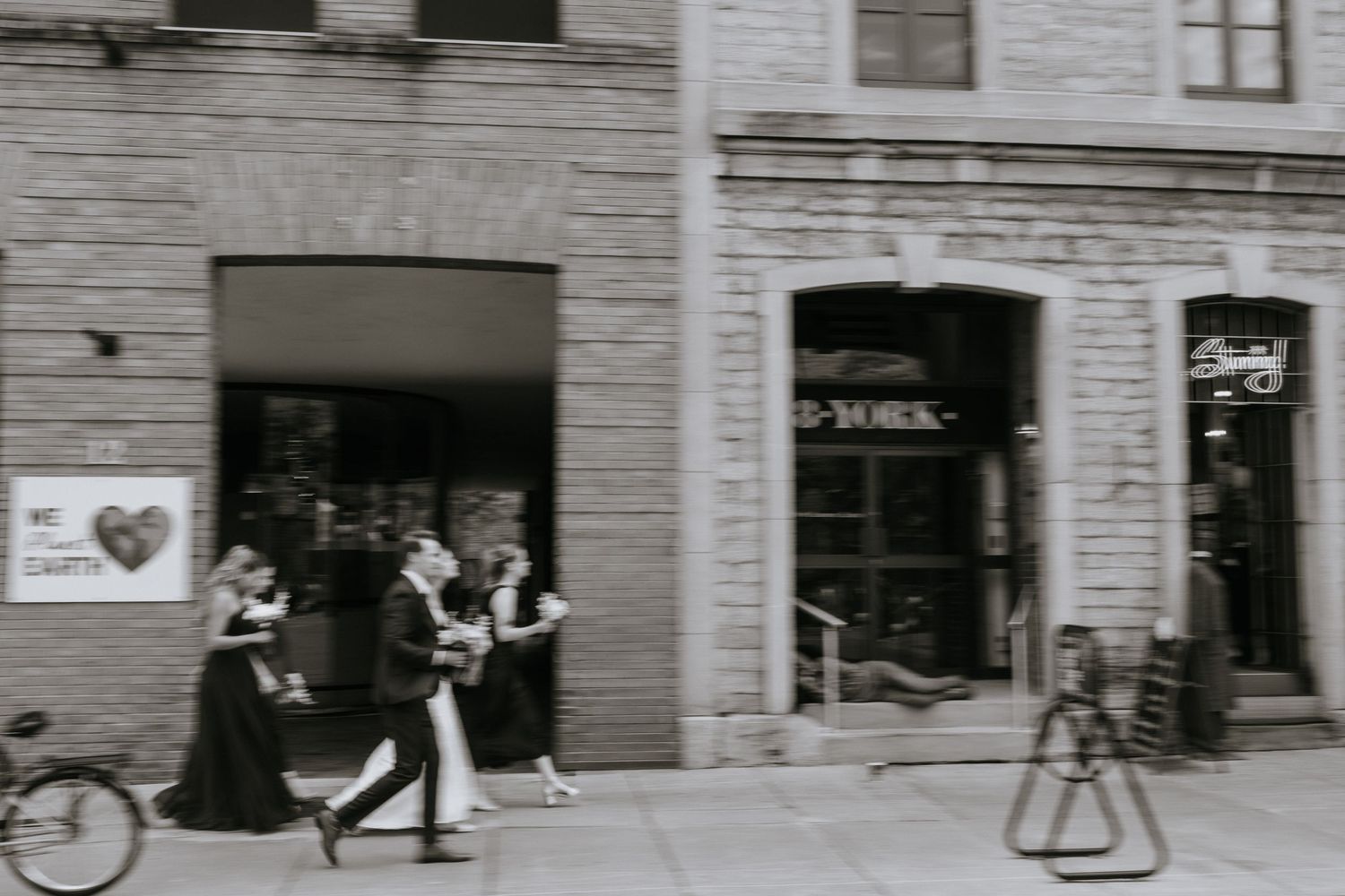 A black and white historical street scene shows storefronts and pedestrians along a city sidewalk in the early 1900s.