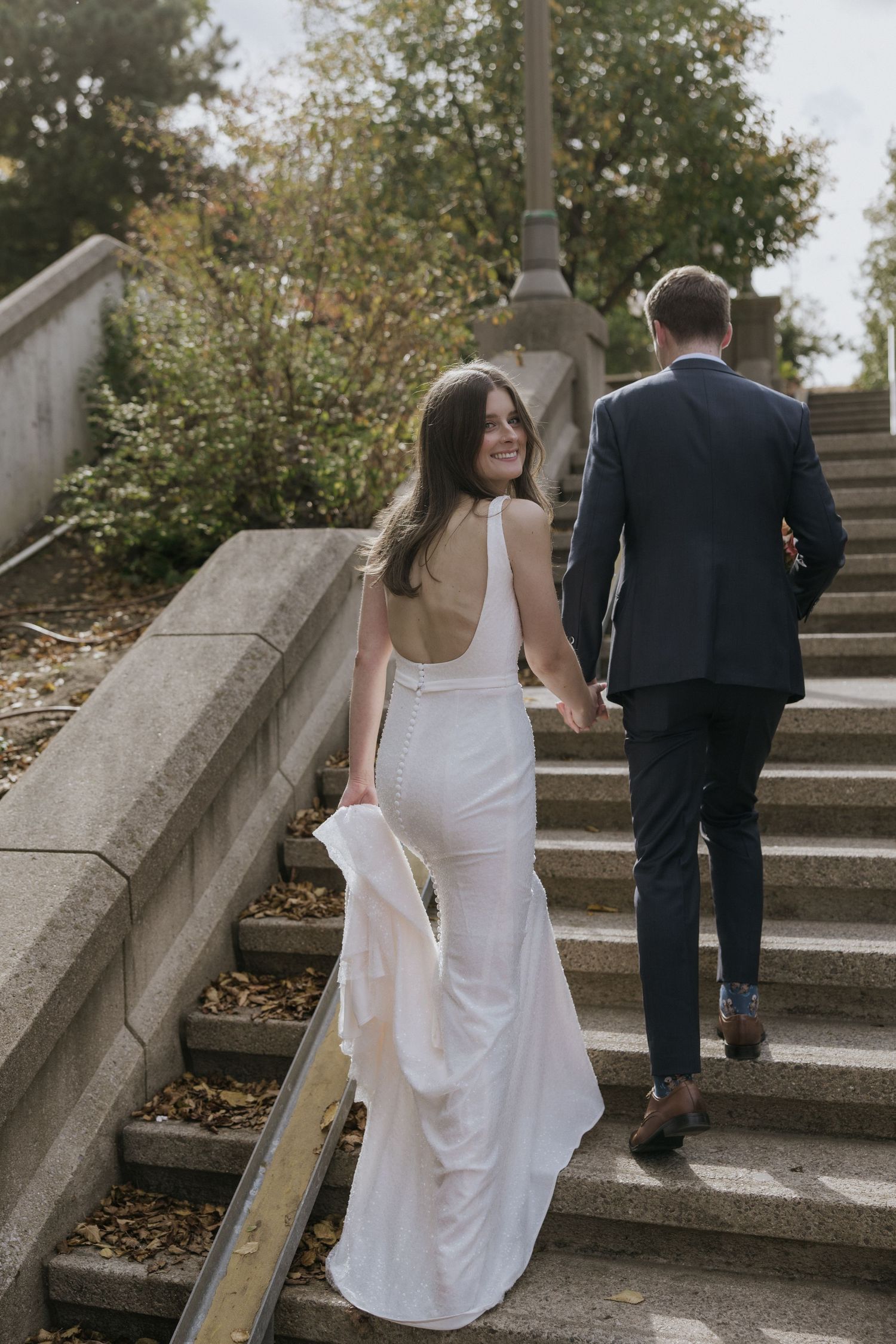 A wedding couple walks up stone garden steps surrounded by autumn foliage and natural light.