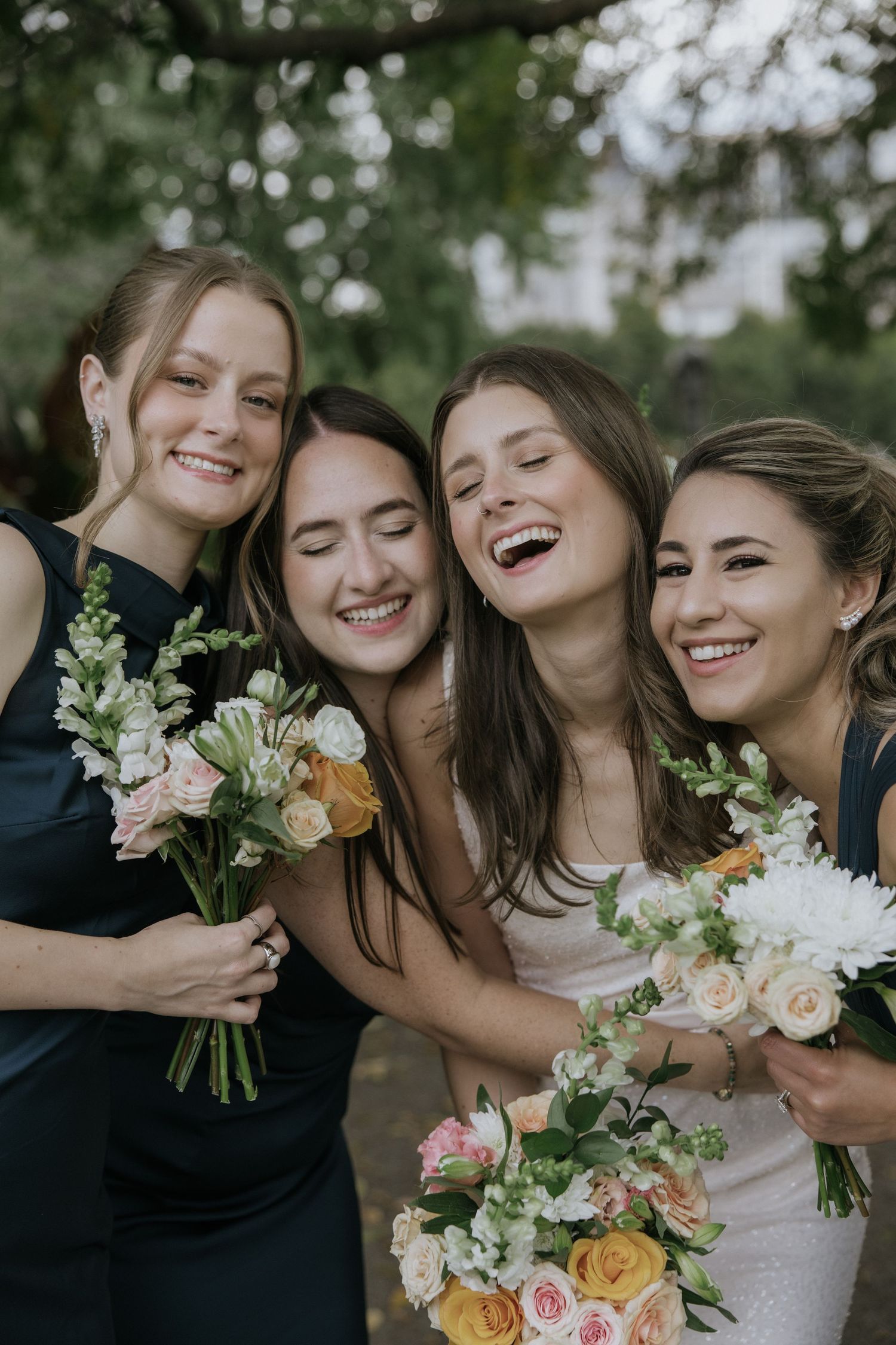 A joyful wedding party of bridesmaids in black dresses hold beautiful bouquets of mixed flowers against a green backdrop.