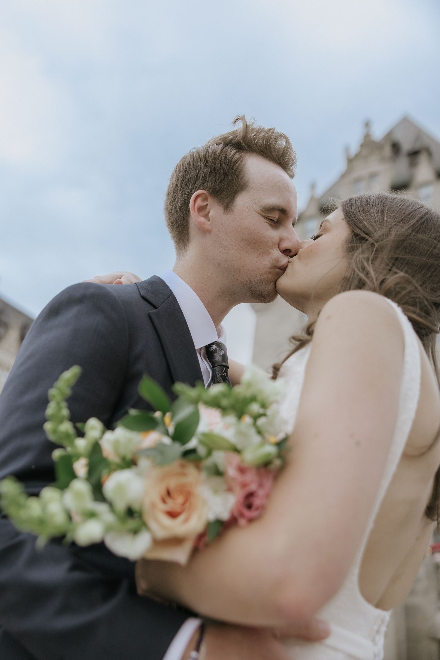 A romantic wedding couple shares a tender kiss while holding a bouquet of pink and peach roses with greenery.