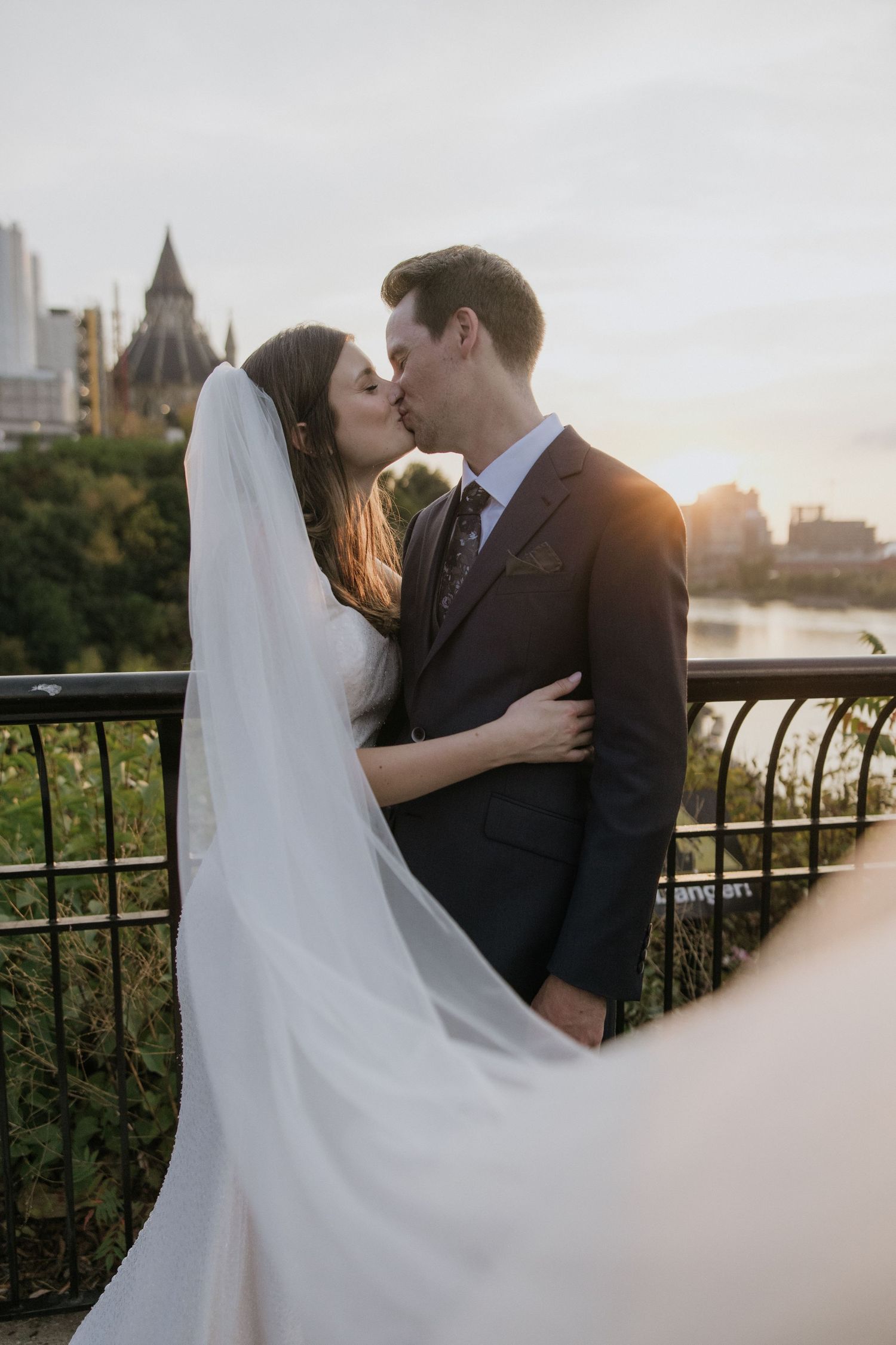 A romantic wedding couple embraces during sunset at a scenic overlook with city buildings visible in the background.