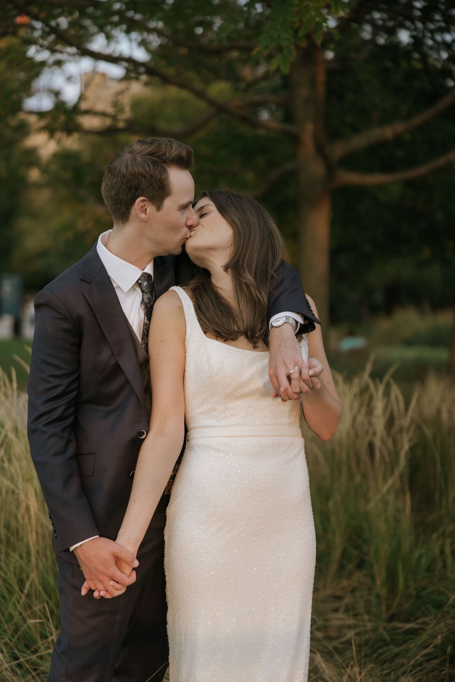 A romantic wedding couple shares intimate kisses in a field of tall grass at sunset.