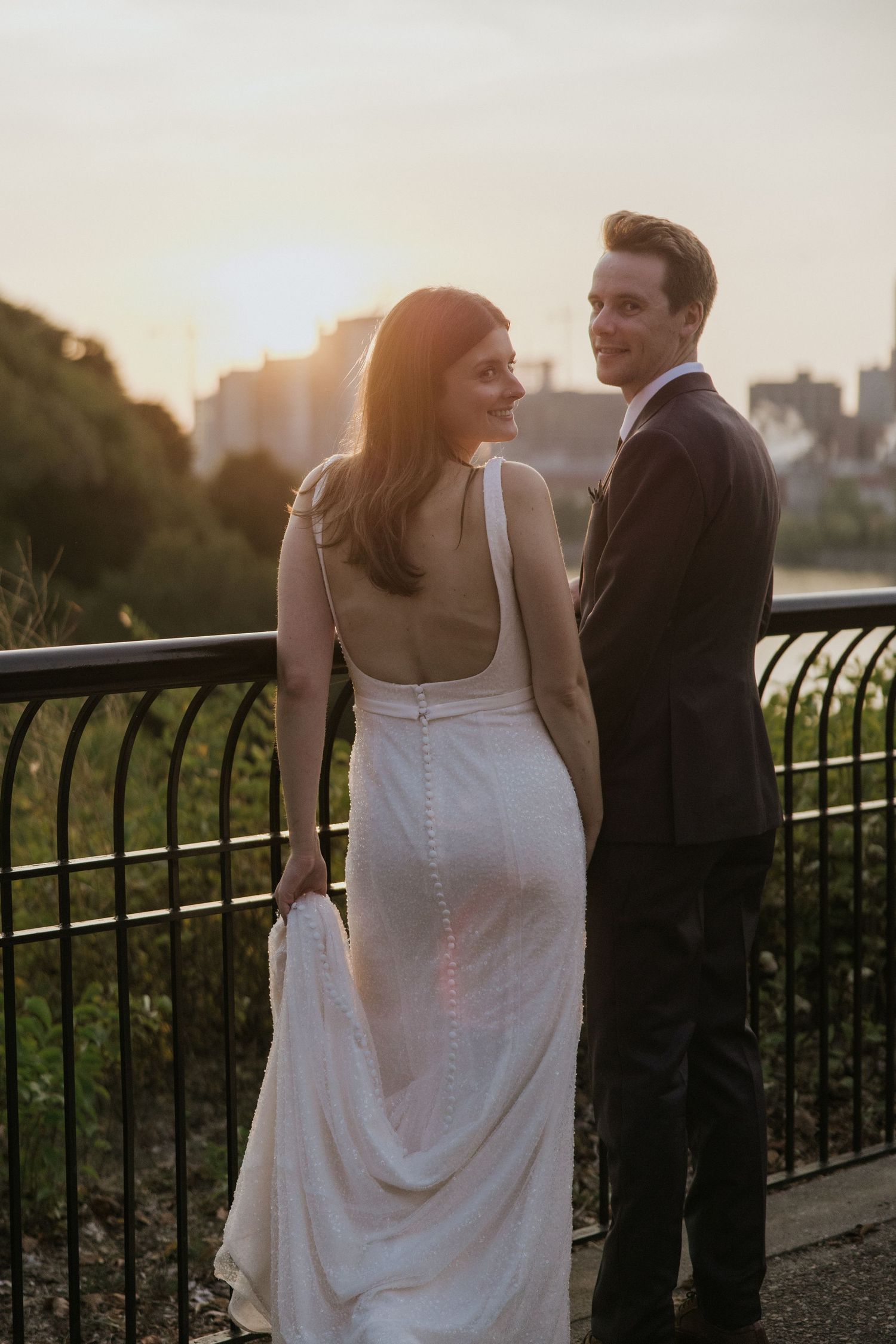 A couple in formal attire stands together on a balcony overlooking a city skyline at sunset.