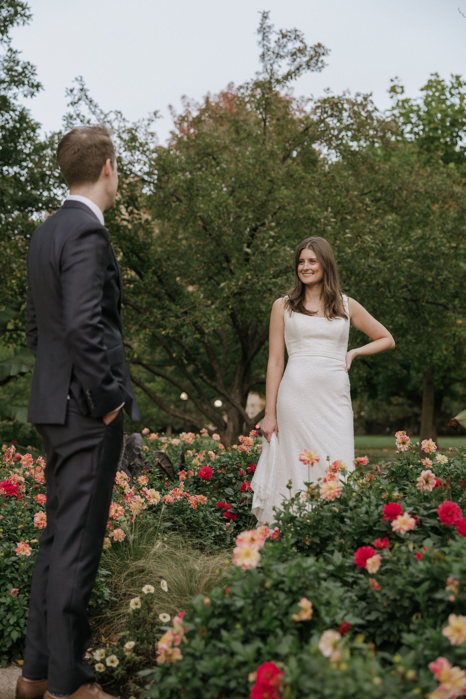 Couple in wedding attire poses romantically among blooming rose bushes in a garden setting.
