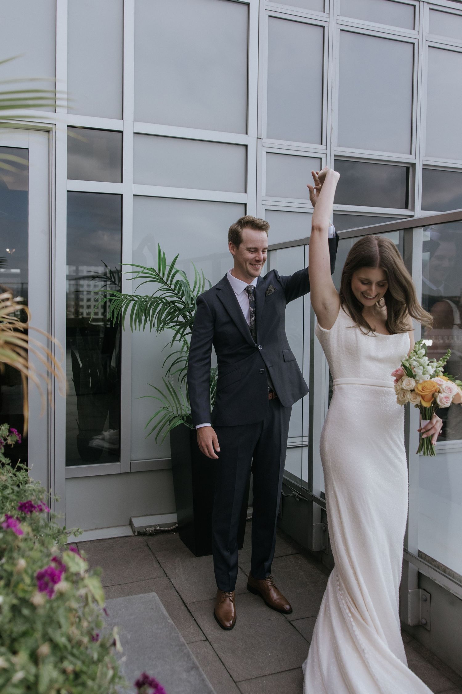 A newlywed couple celebrates joyfully on a terrace with glass windows and potted plants in the background.