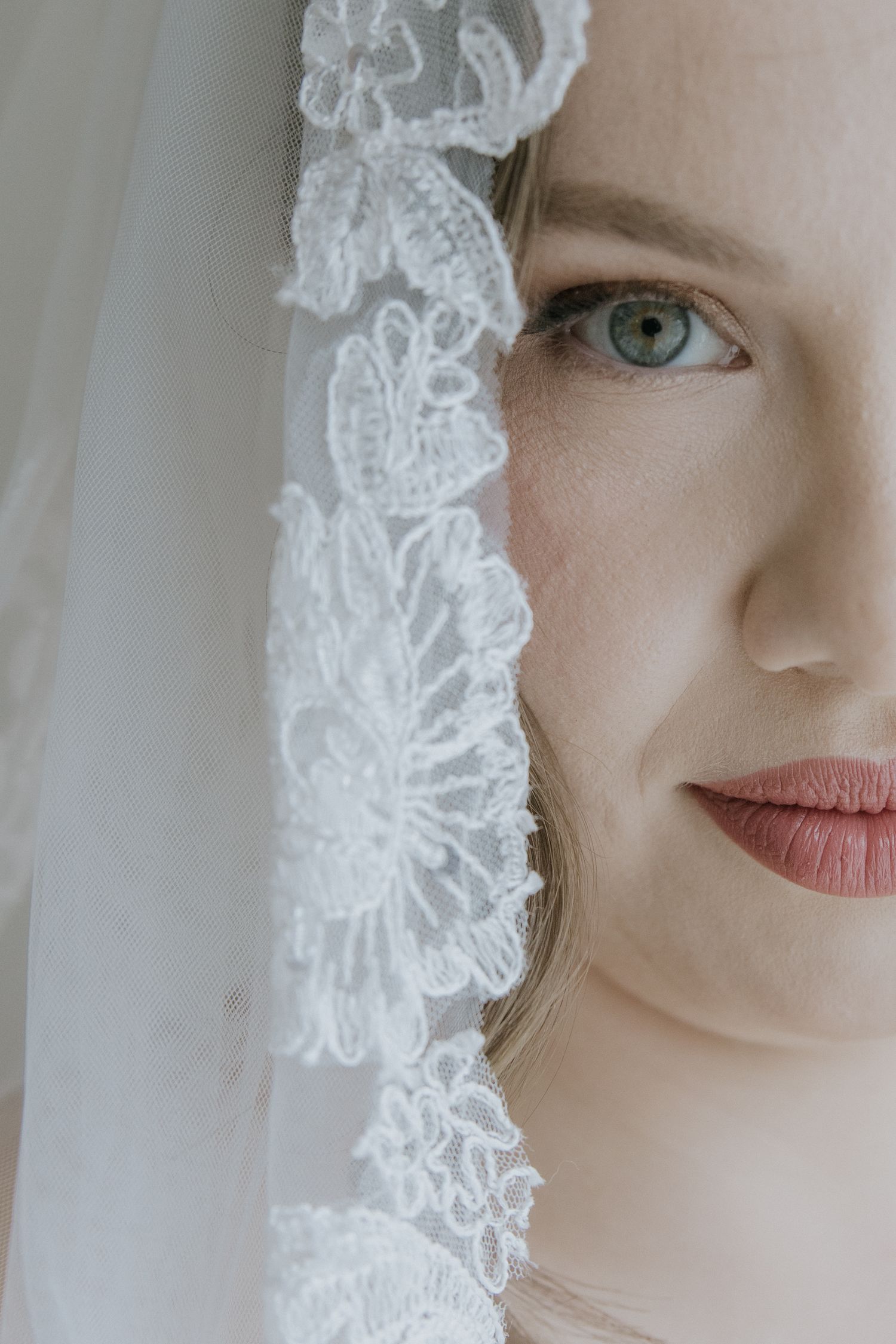 Close up portrait behind delicate white lace veil with floral pattern showing half of face and bright blue eye.