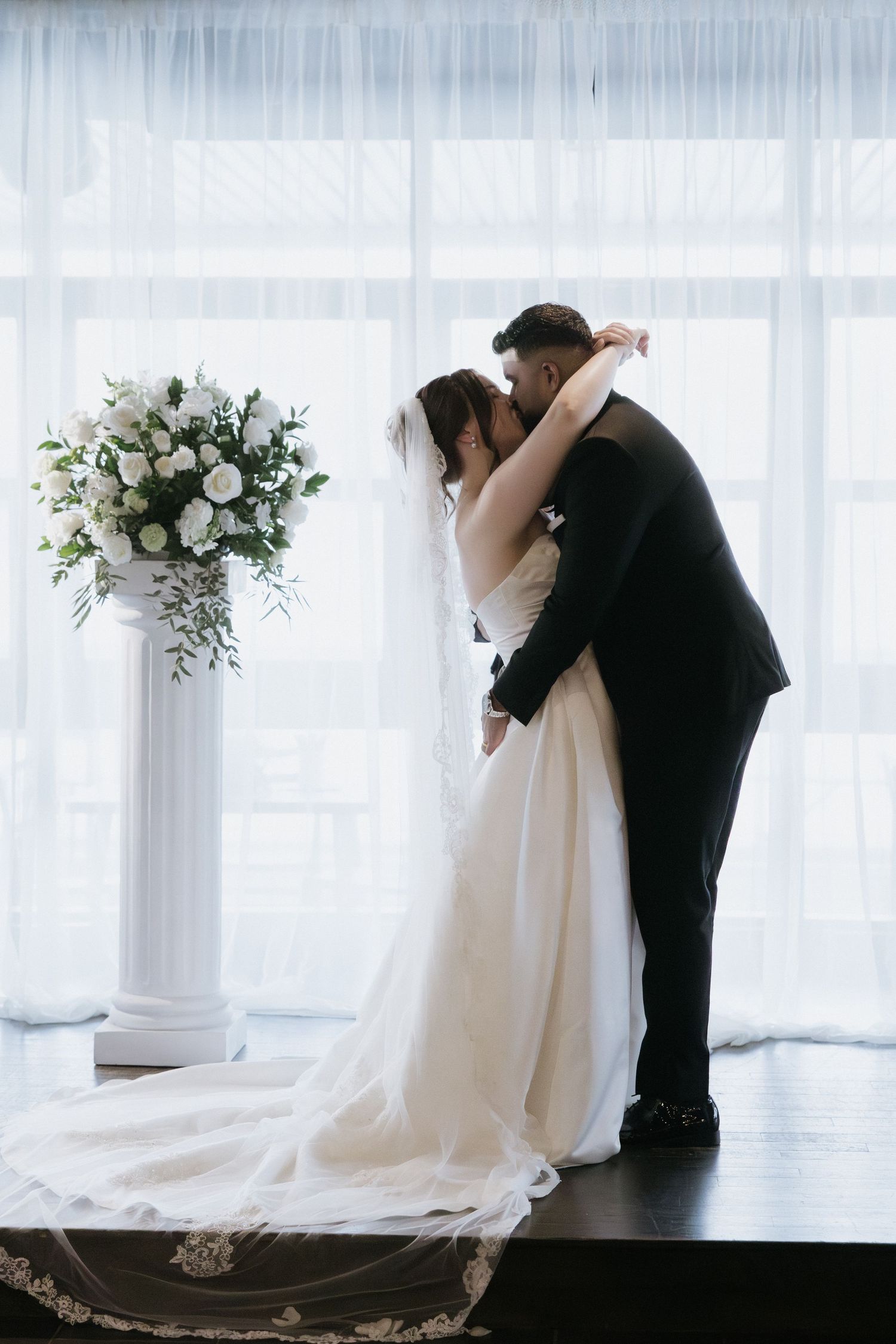 Three romantic wedding photos show a couple embracing in a light-filled room with white floral arrangements.
