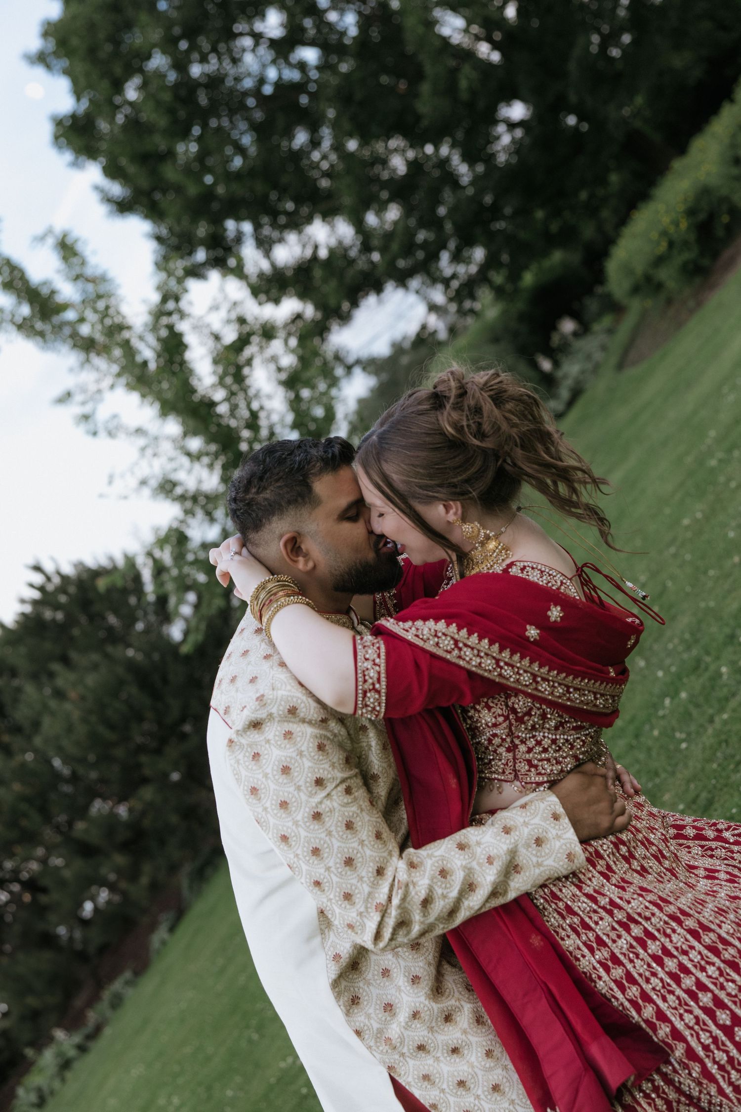 A couple in traditional wedding attire embrace and dance outdoors, with the bride wearing a red and gold lehenga.