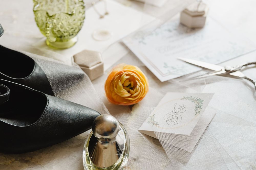 Black leather flats, wedding stationery, and a yellow ranunculus flower arranged on a white marble surface with glassware.