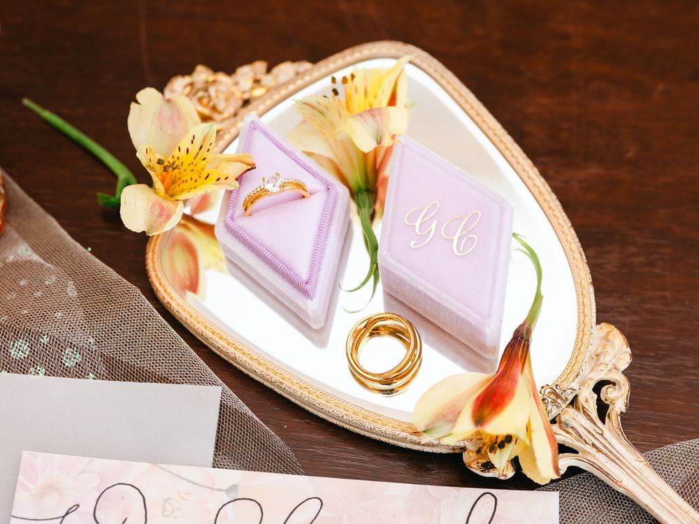 Ornate vintage mirror reflecting pink wedding invitations, yellow lilies, and golden wedding rings.