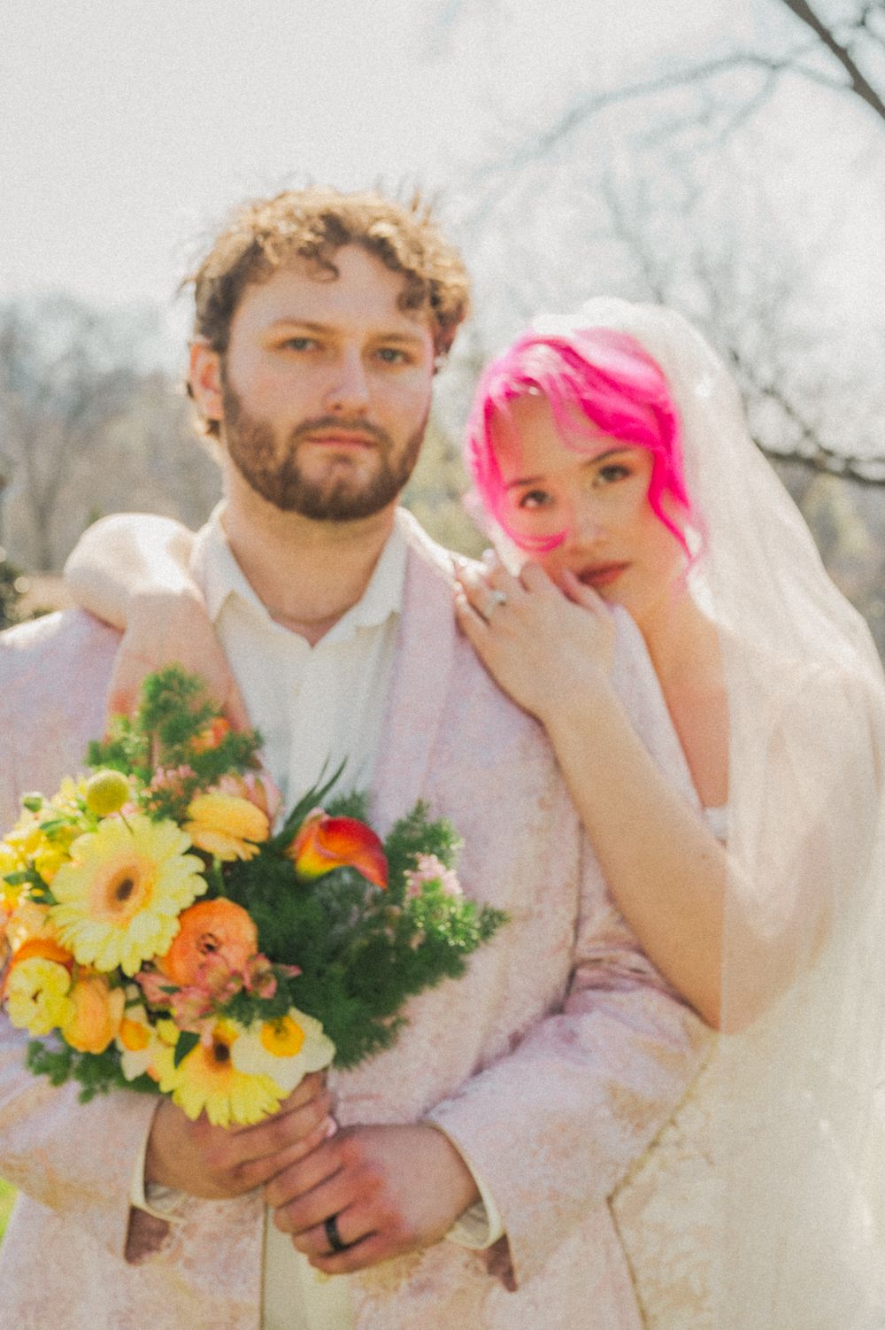 A wedding couple stands outdoors with the bride wearing a veil and holding a vibrant yellow and orange floral bouquet.
