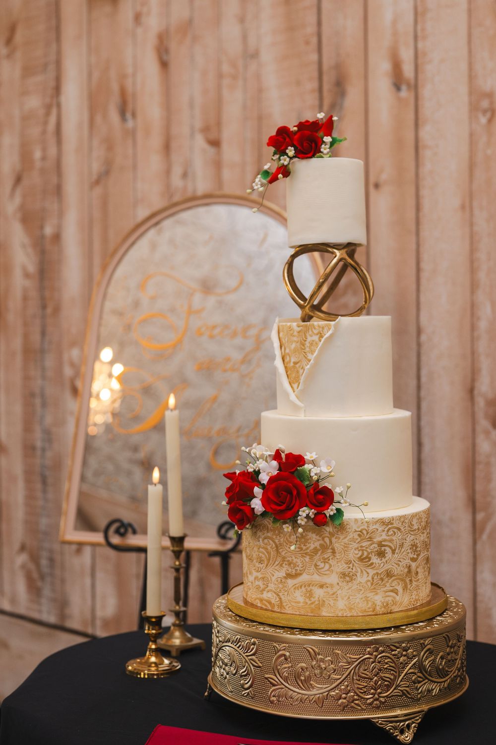 Elegant white wedding cake with gold geometric details and red rose accents displayed beside lit candles on a black table.