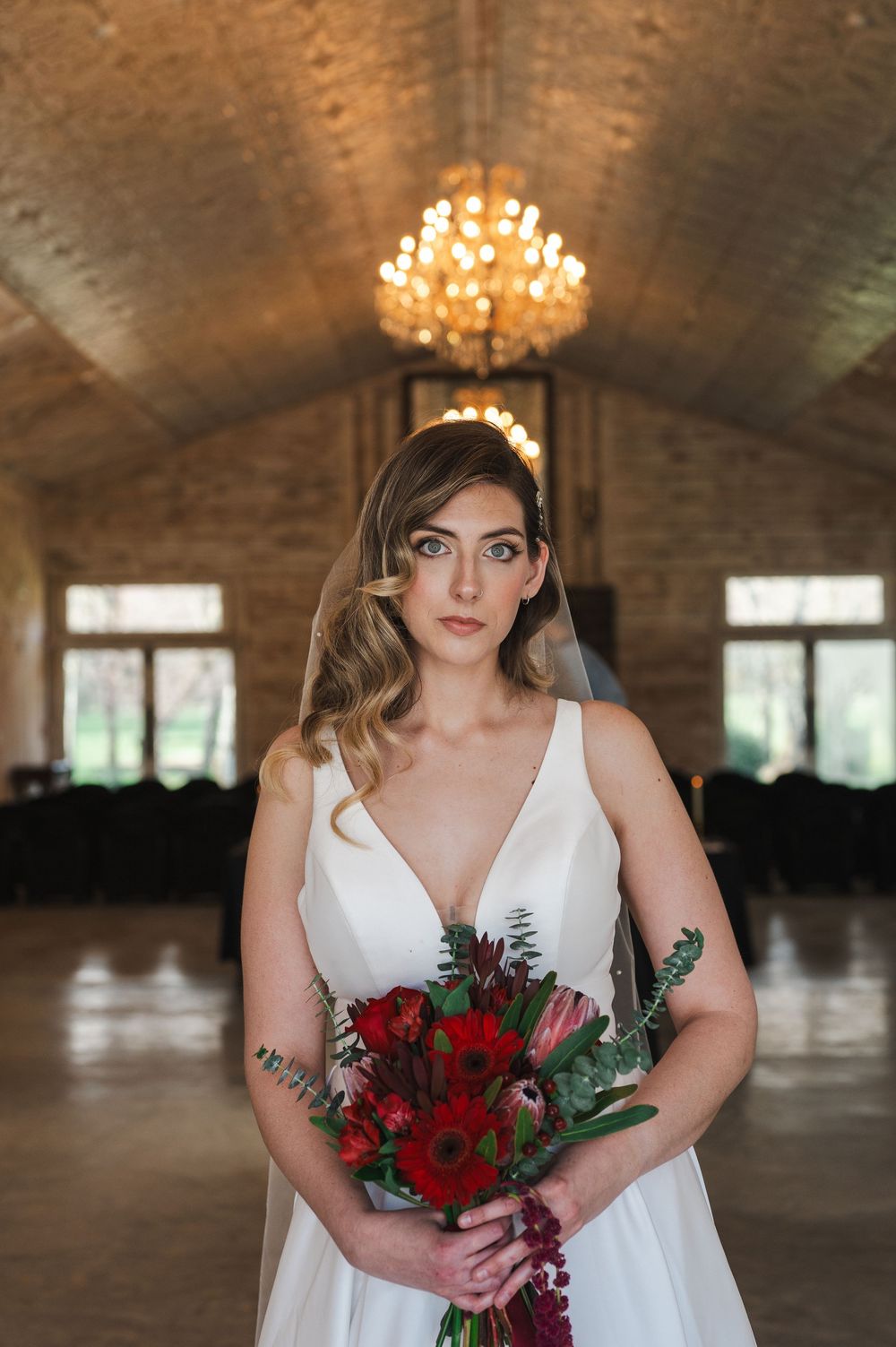 Elegant bride in white v-neck wedding dress holds vibrant red bouquet in grand venue with chandelier and arched ceiling.