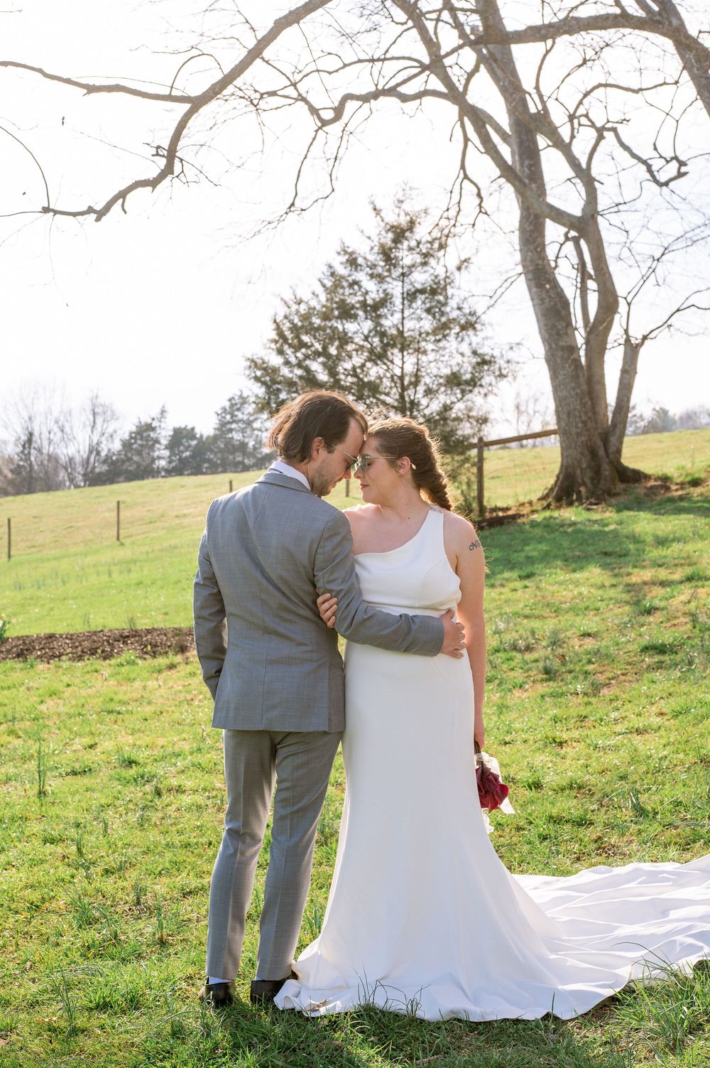 A romantic wedding couple stands together in a springtime meadow under a large tree on a sunny afternoon.