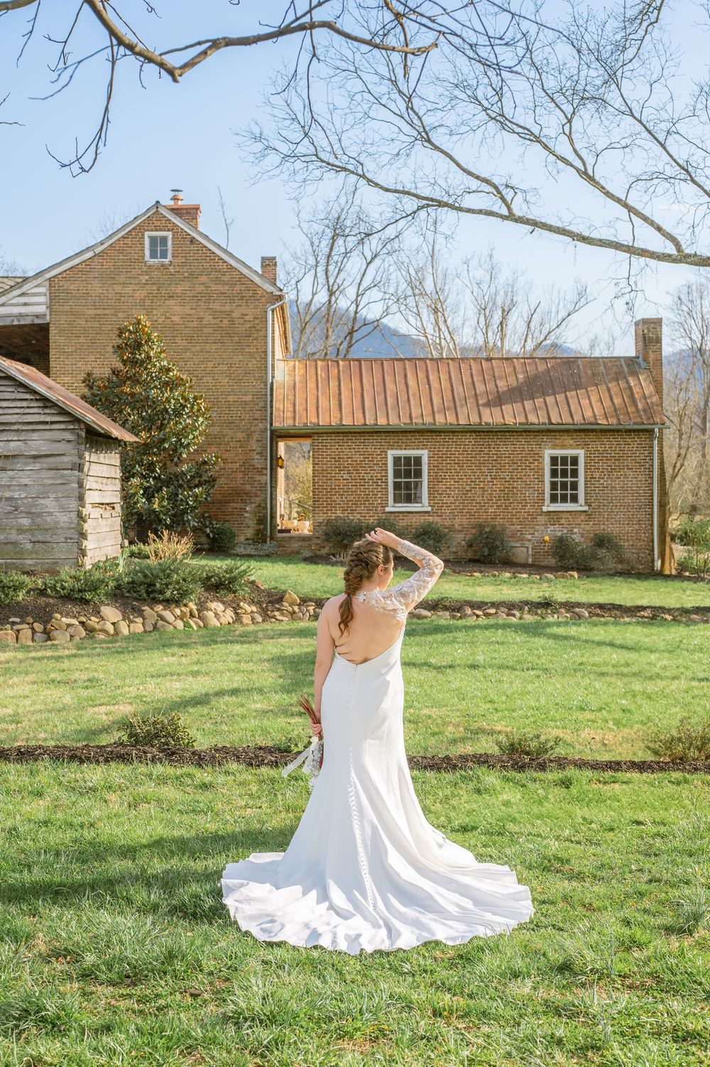 A flowing white wedding dress catches sunlight on a grassy lawn outside a historic brick farmhouse on a sunny day.