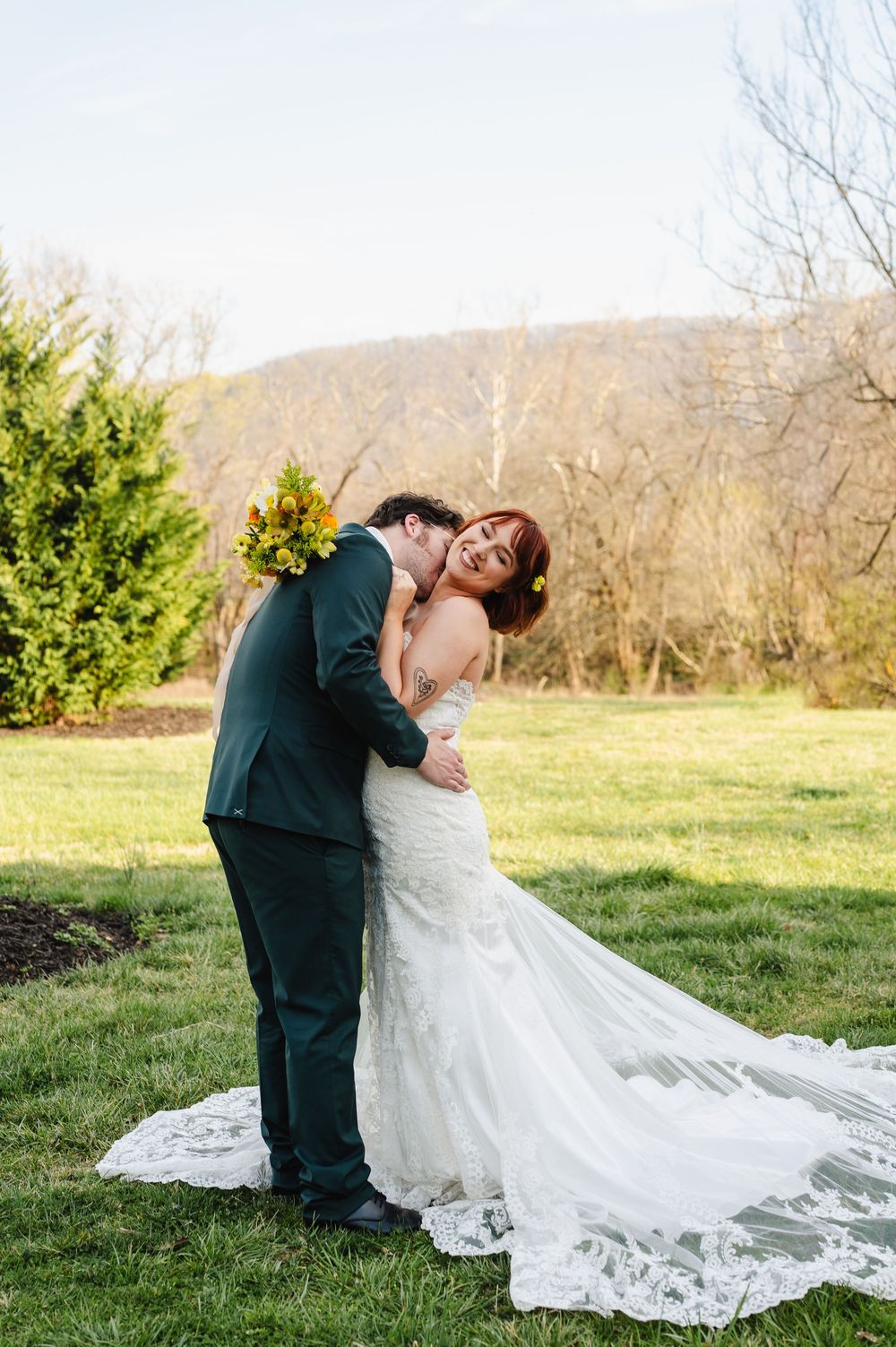 A romantic outdoor wedding moment with a mountainous backdrop as a couple embraces on a grassy lawn.