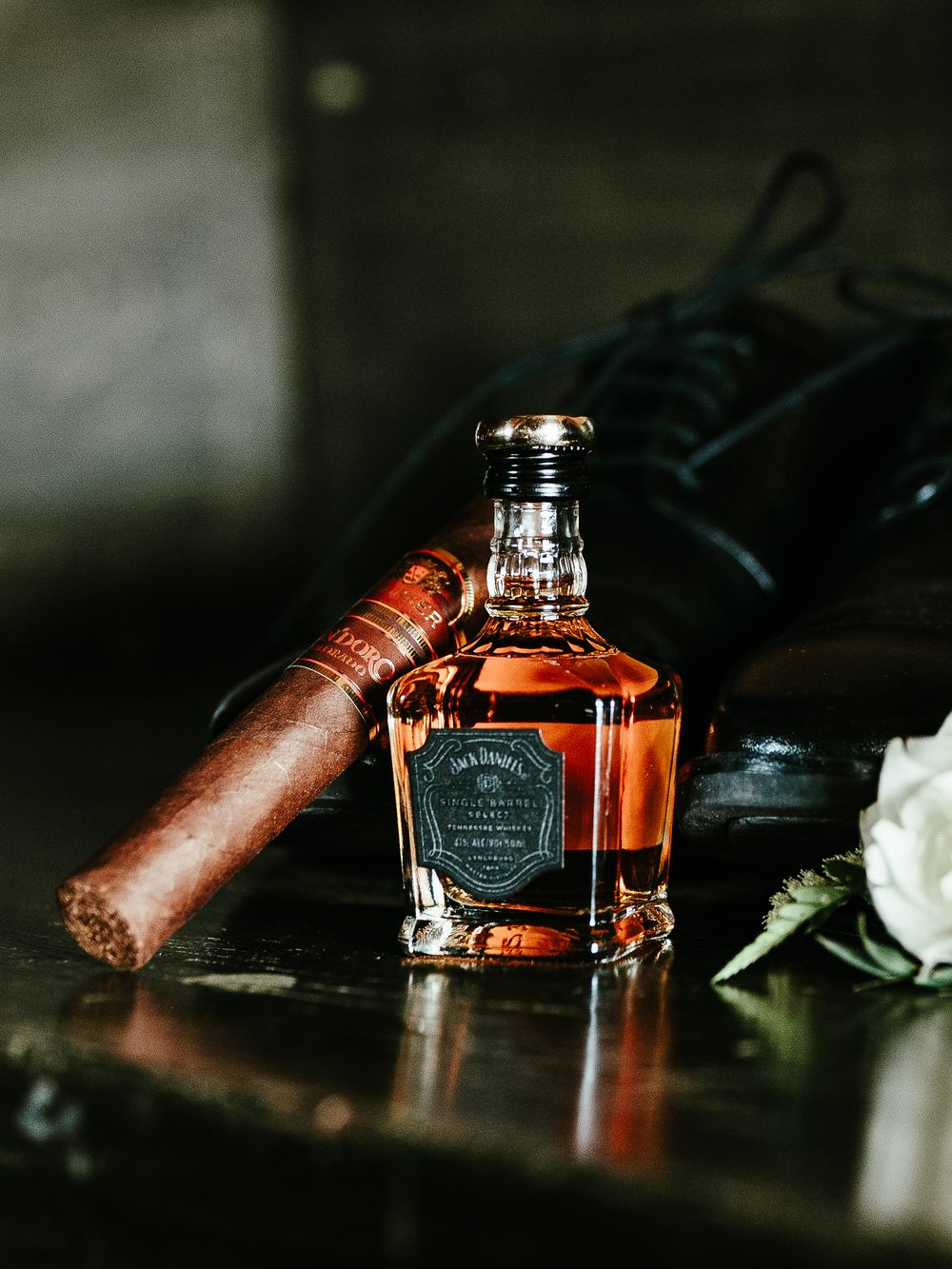 A bottle of premium whiskey alongside a lit cigar and black leather shoe in a moody, dark setting.