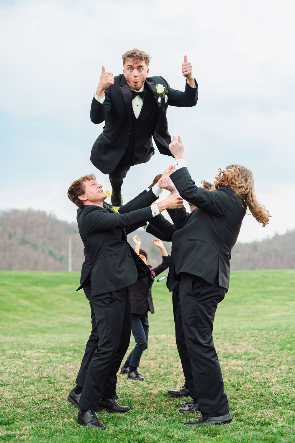 Groomsmen in black tuxedos playfully lift and toss one member of the wedding party on a grassy hillside.