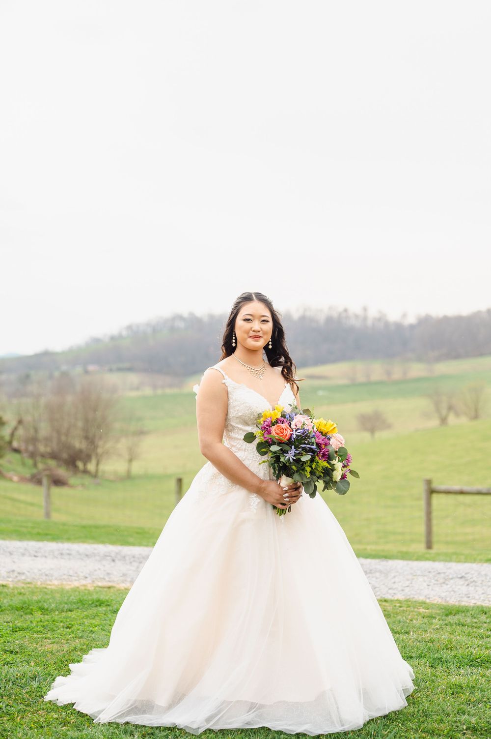 A bride in a flowy white ballgown holds a colorful bouquet while standing on green countryside grounds.