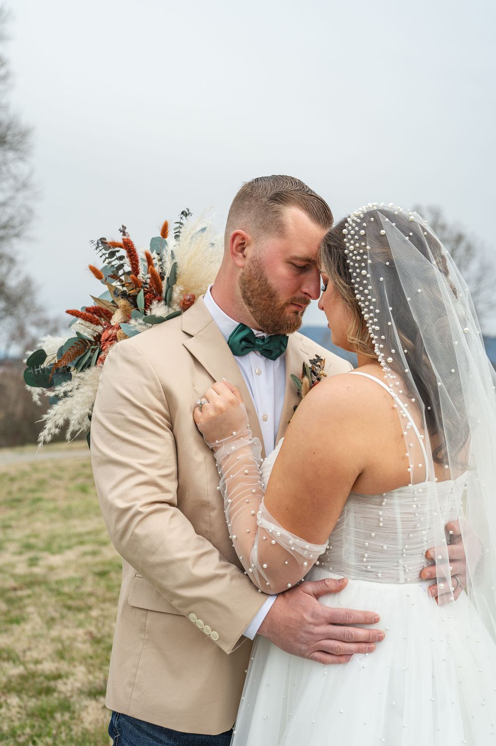 A romantic wedding couple embracing in a field wearing beige suit and white dress with floral details.