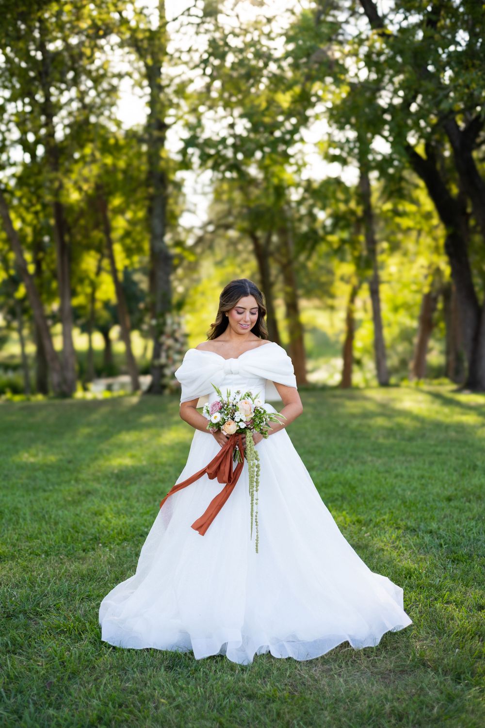 A bride in an off-shoulder white wedding gown holds a bouquet while standing in a sunlit garden setting.