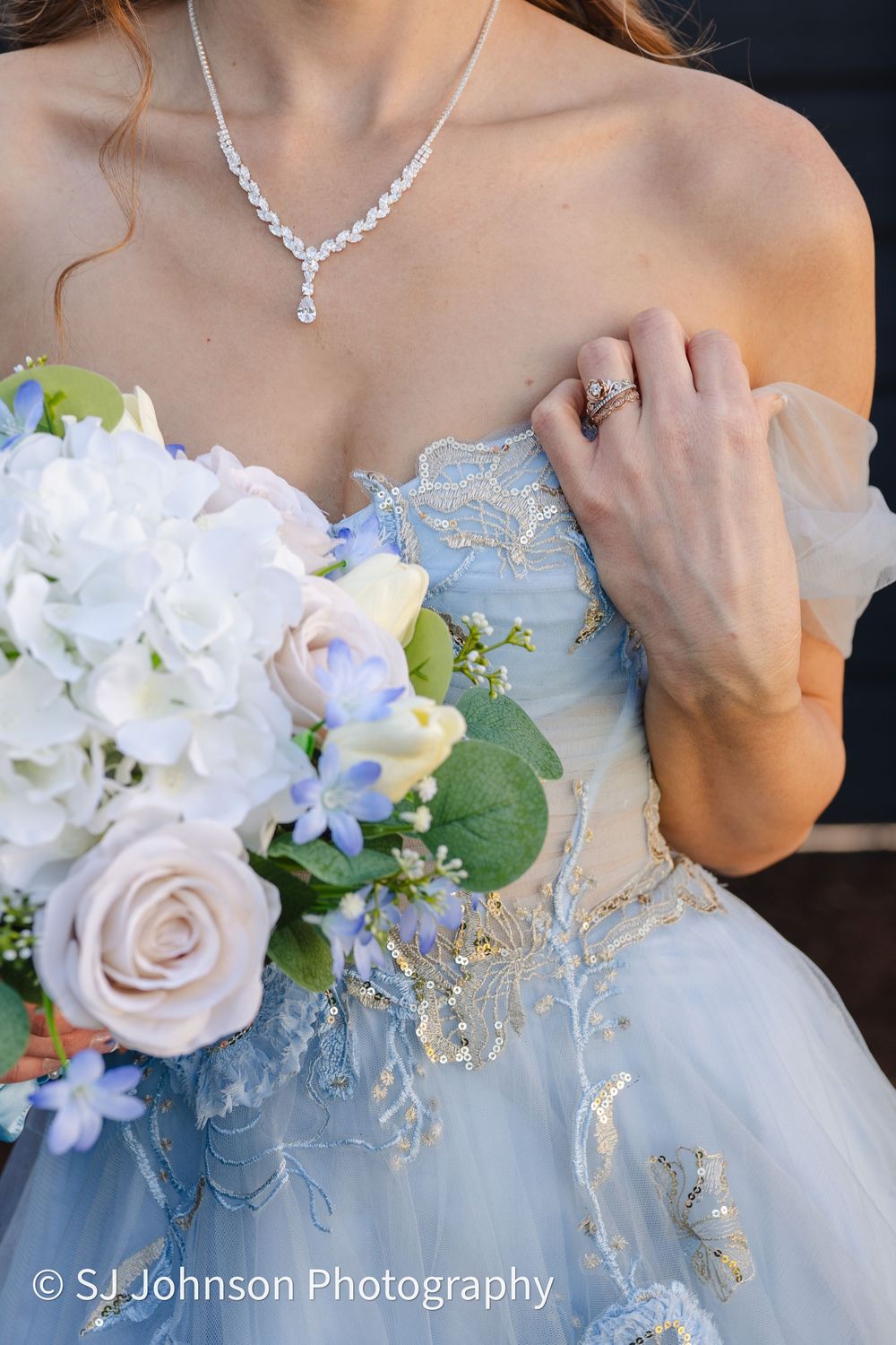 Close-up of blue wedding gown with lace details, delicate crystal necklace, and white and blue bridal bouquet.