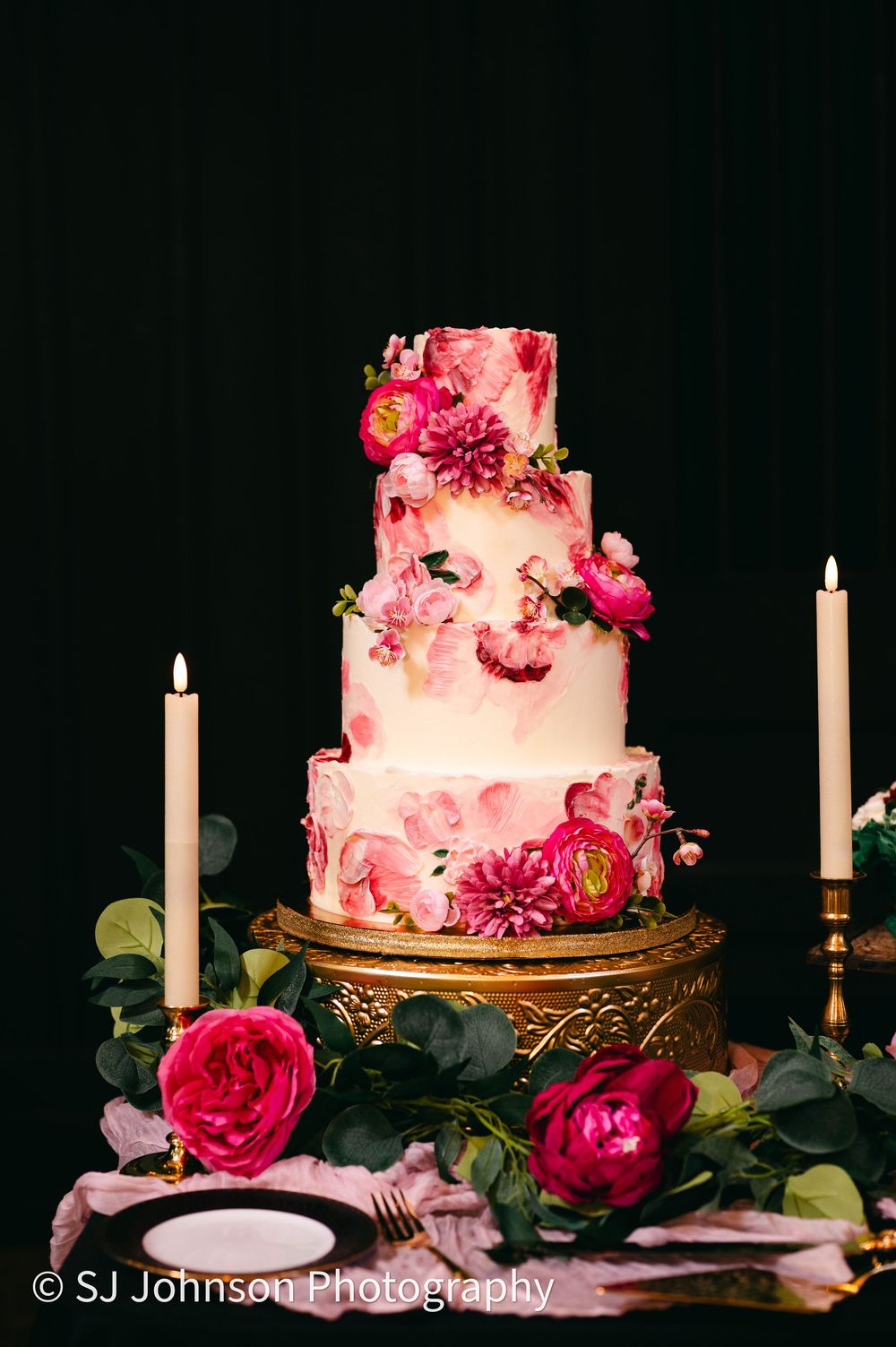 Elegant four-tier pink wedding cake decorated with fresh flowers on ornate gold stand with white candles and dark background.