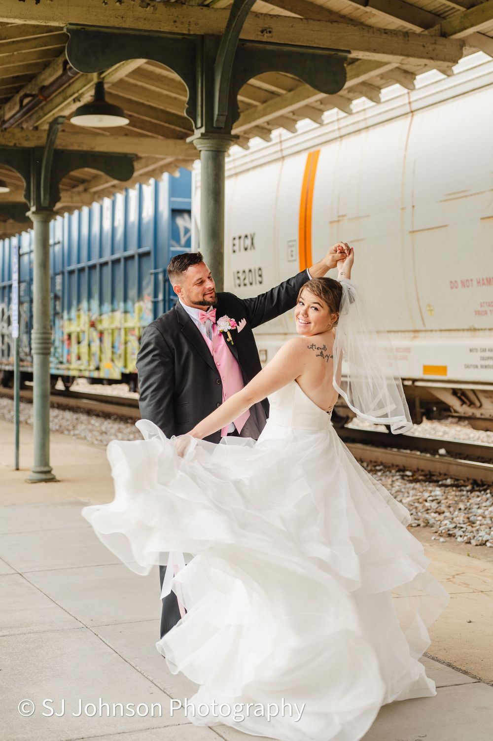 A bride in a flowing white gown dances with her groom at a vintage train station platform.
