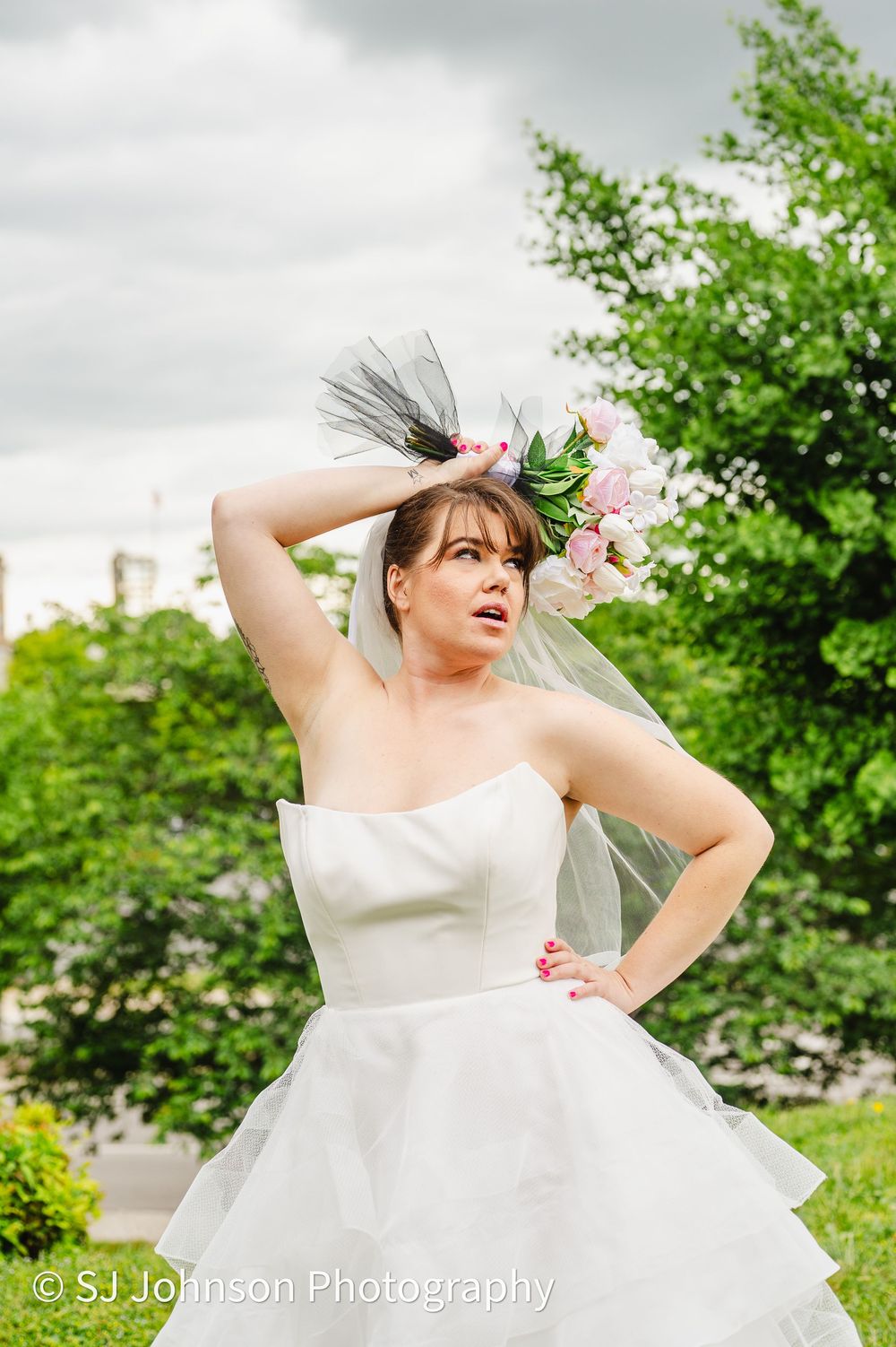 A bride in a white strapless wedding gown with tulle skirt poses outdoors among greenery on an overcast day.