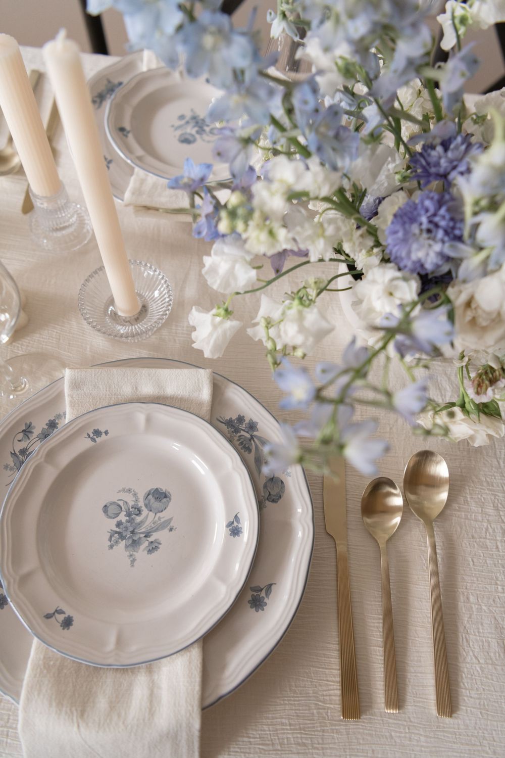 Vintage china place setting with blue floral pattern and gold utensils beside delicate flowers.