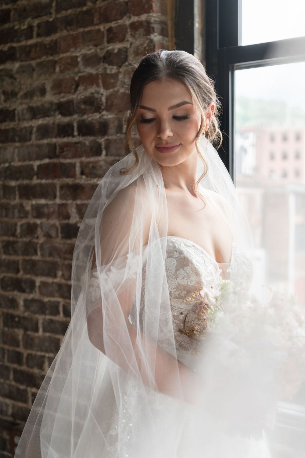 Ethereal bridal portrait by brick window with flowing veil and lace gown.