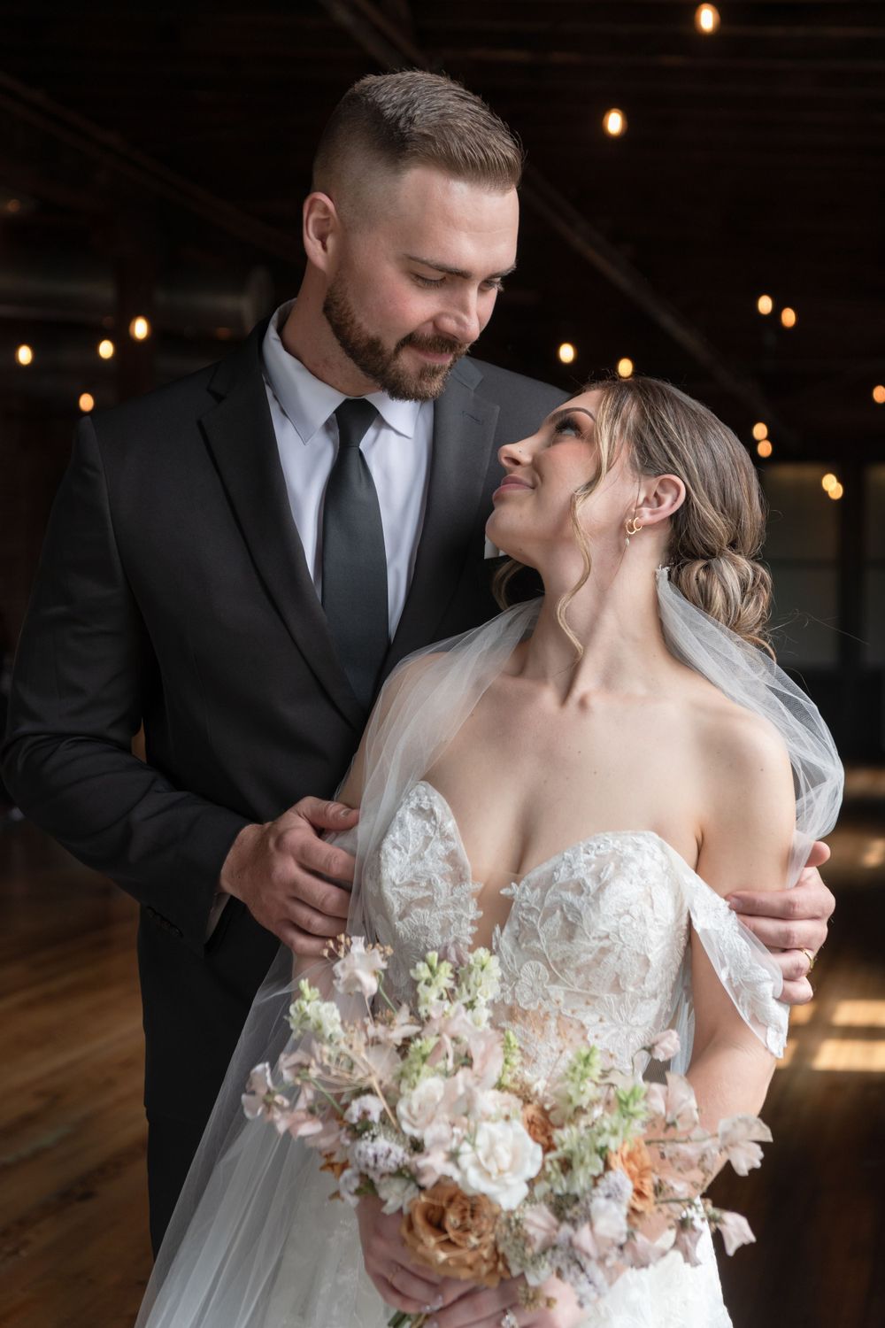 Intimate wedding portrait in industrial venue with string lights and natural bouquet.