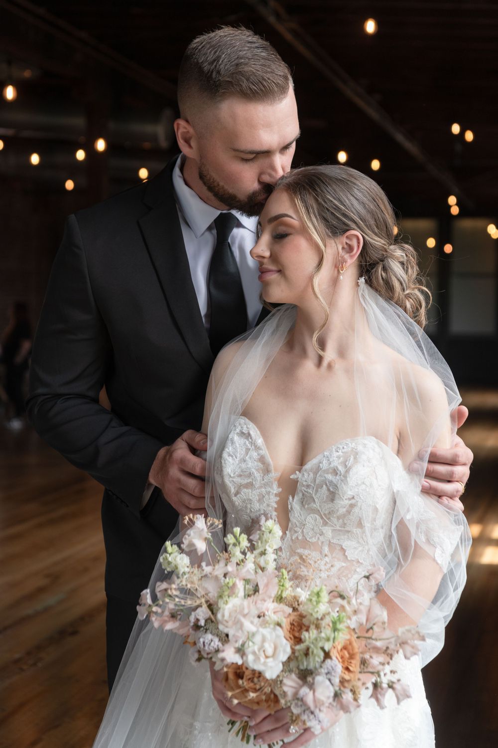 Tender wedding moment captured in industrial space with string lights overhead.