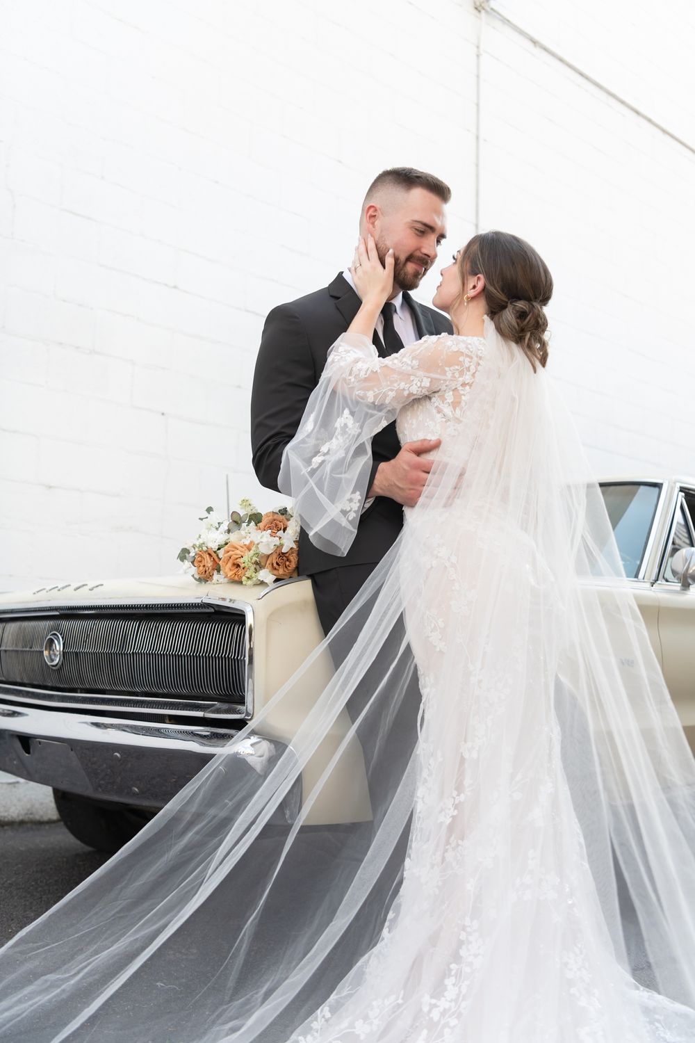 A romantic wedding couple embraces next to a vintage car with a flowing white veil.