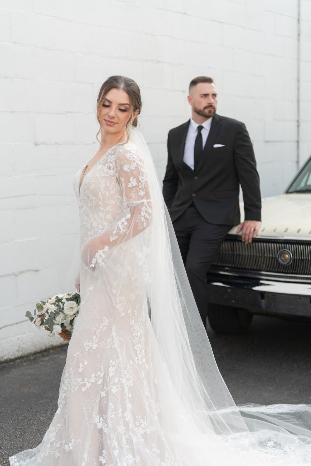 Elegant couple poses beside vintage car, bride wearing lace gown with cathedral veil.