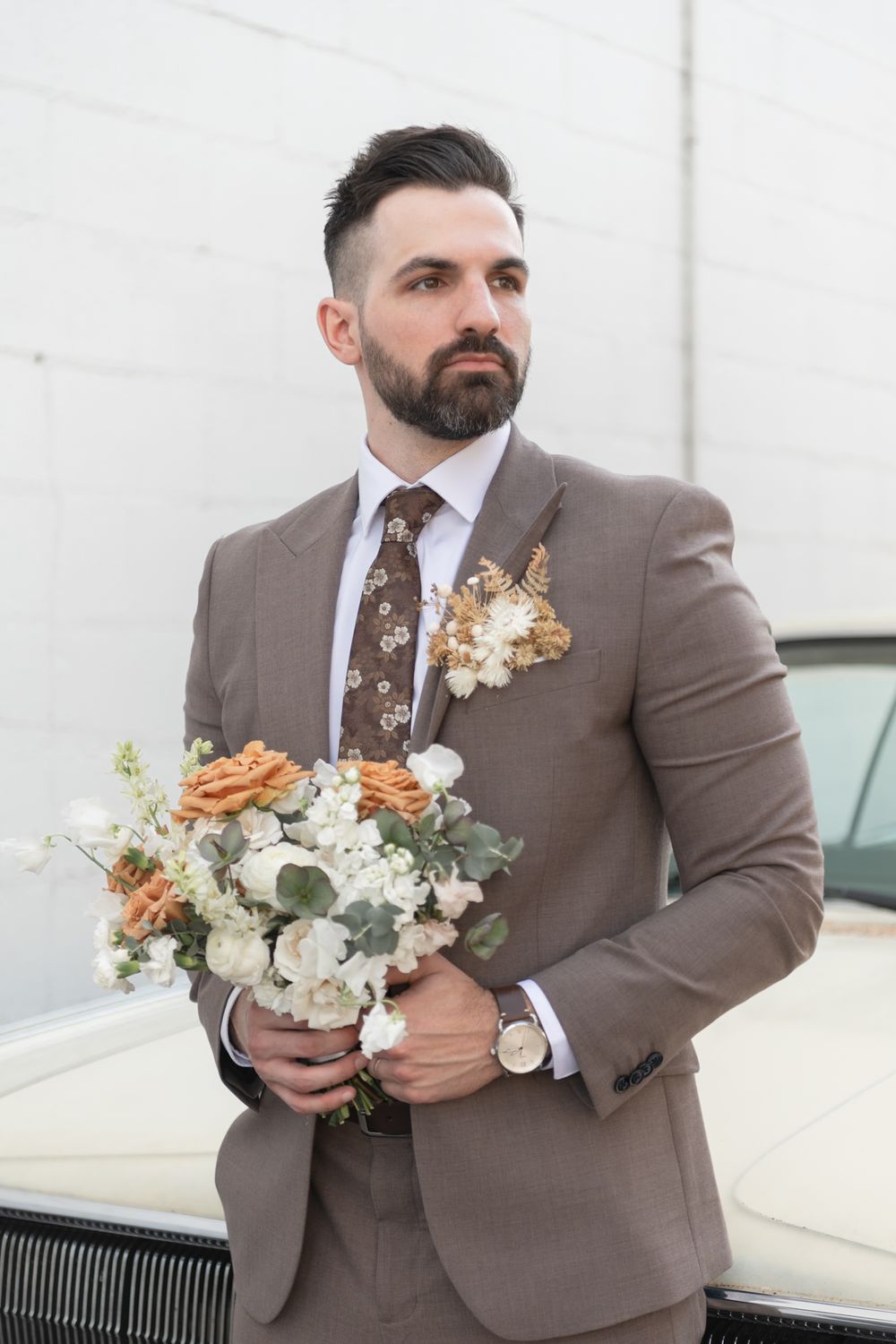 Groom in brown suit holds romantic bouquet of white and peach flowers against white wall.