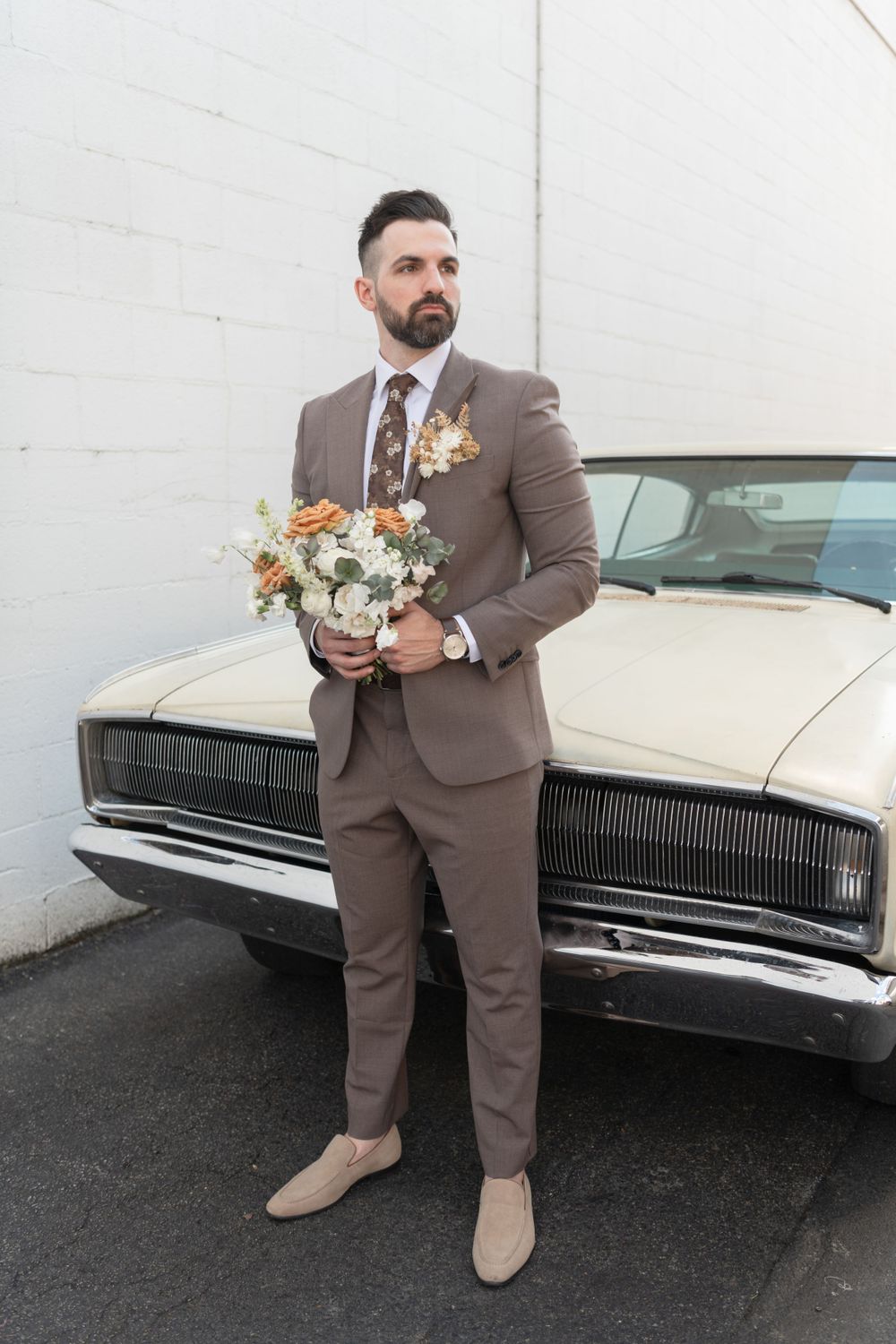 A groom in a taupe suit holds a floral bouquet while standing next to a vintage white car.