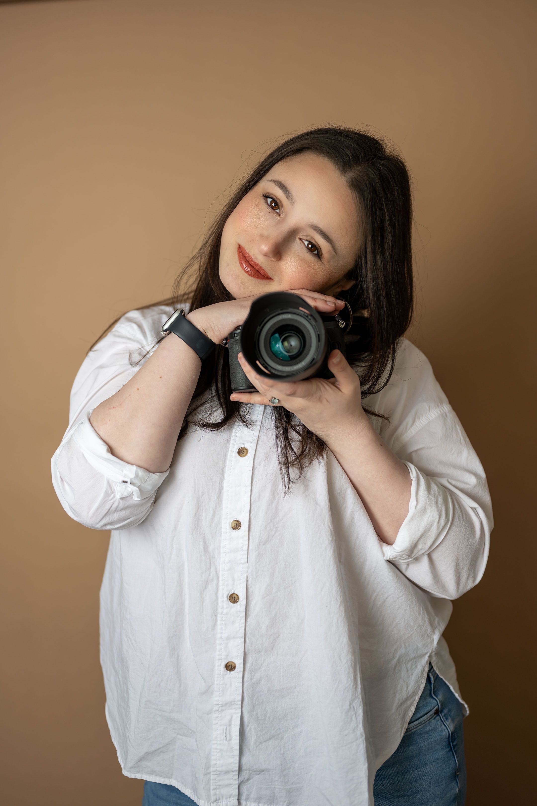 Smiling woman in white shirt holding DSLR camera close to face on beige background.