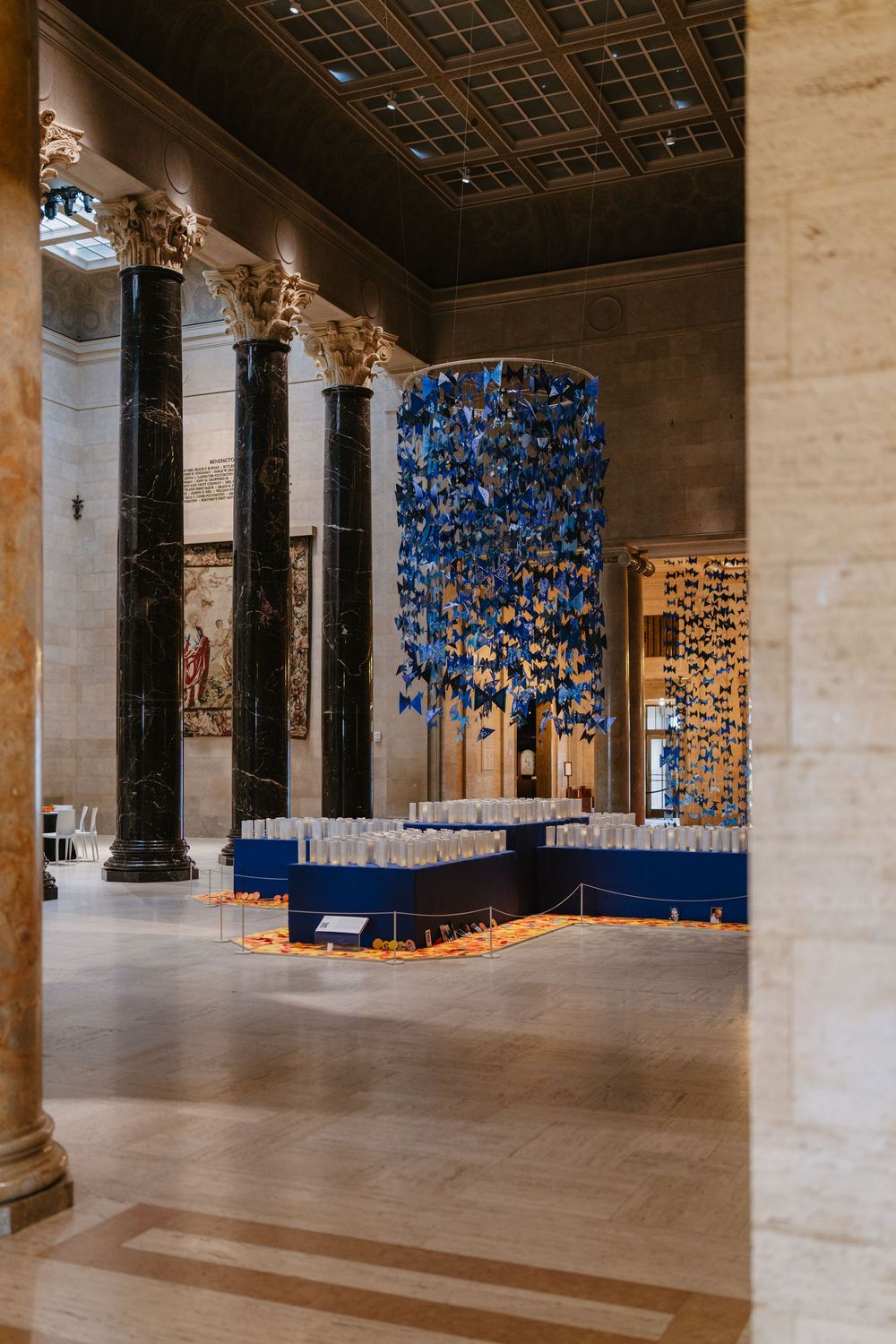 Grand museum hall with blue butterfly art installation suspended from ceiling, flanked by ornate marble columns.