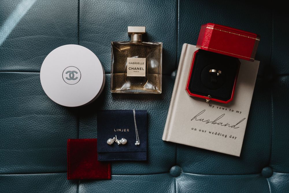 Chanel perfume, Cartier ring box, pearl necklace, and wedding book arranged on teal leather surface.