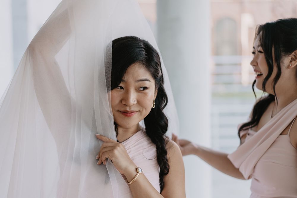 Bridesmaid carefully placing a white veil on a smiling Asian bride in a pink dress during wedding preparations.