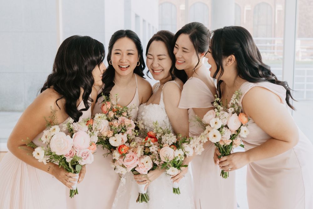 Five bridesmaids in blush dresses laughing together while holding elegant pink and white floral bouquets at a wedding.