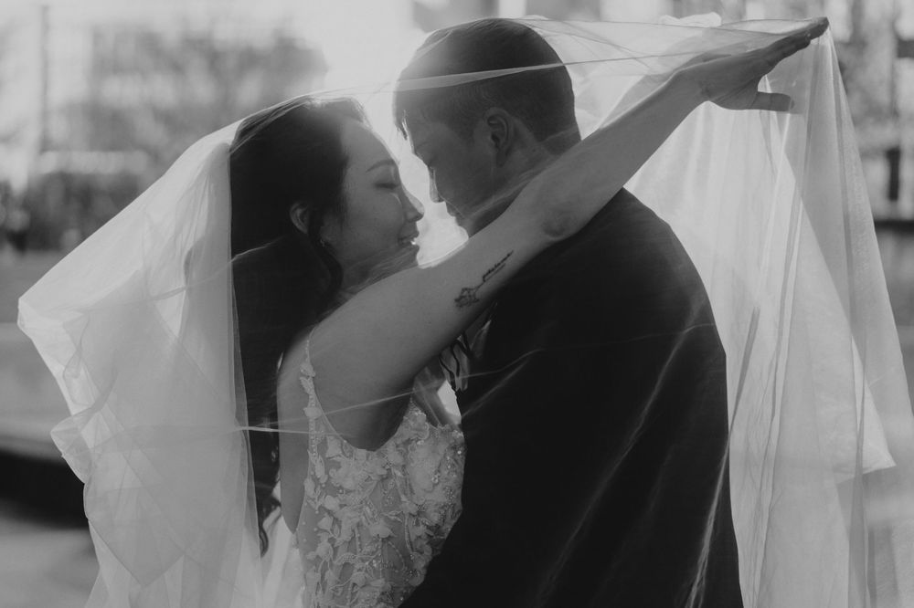 Black and white wedding photo of bride and groom sharing romantic moment under flowing veil.
