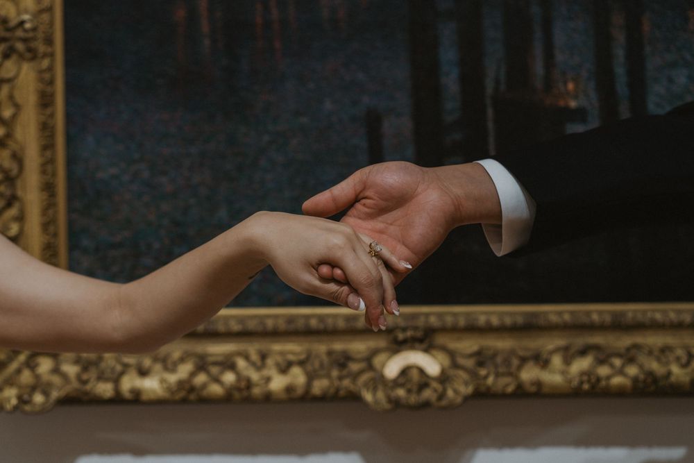 Bride and groom gently holding hands in front of an ornate gold-framed painting on their wedding day.